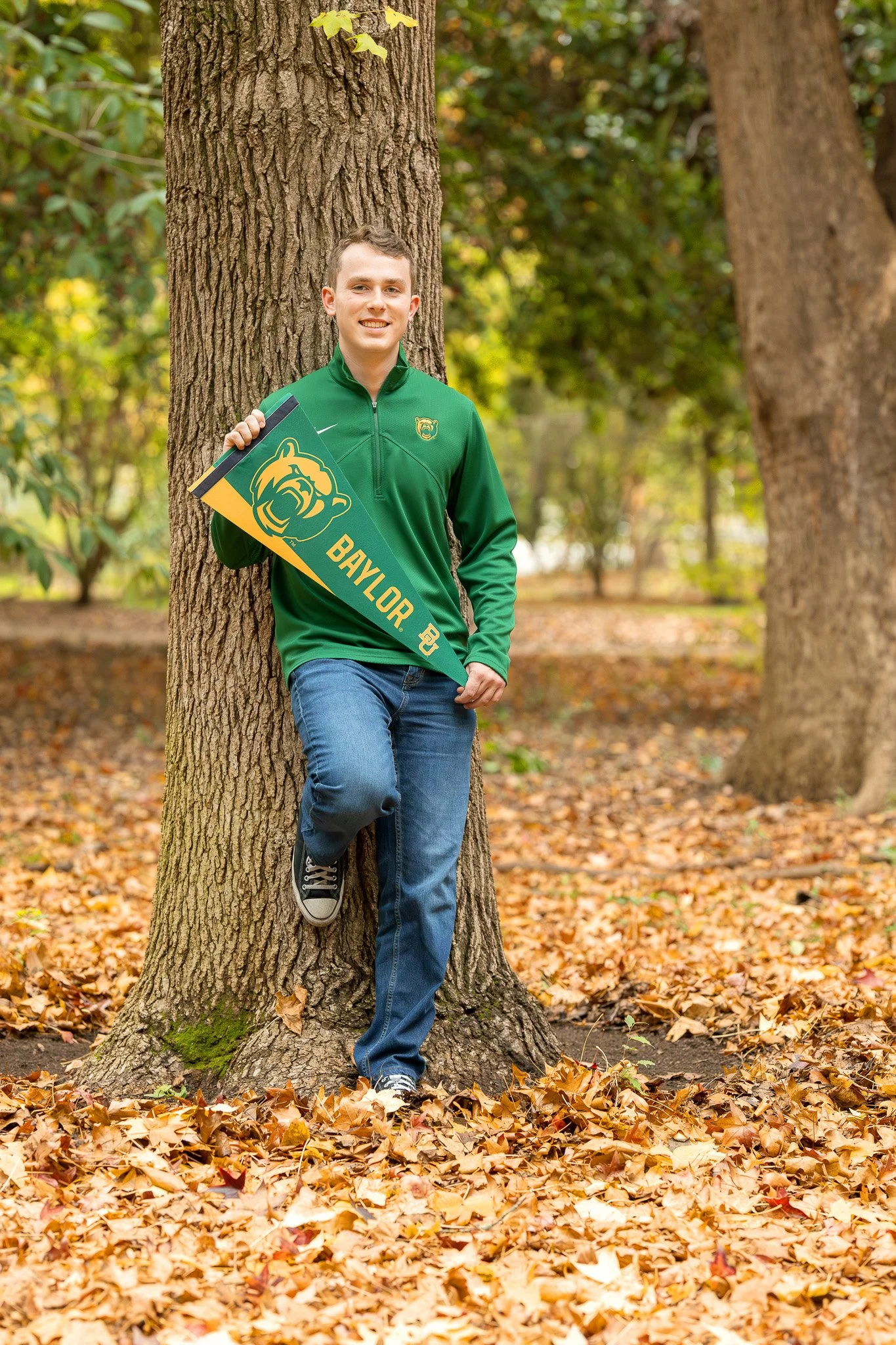 A young man in a green Baylor jacket holding a Baylor pennant leaning against a tree in an autumnal park surrounded by fallen yellow and brown leaves.