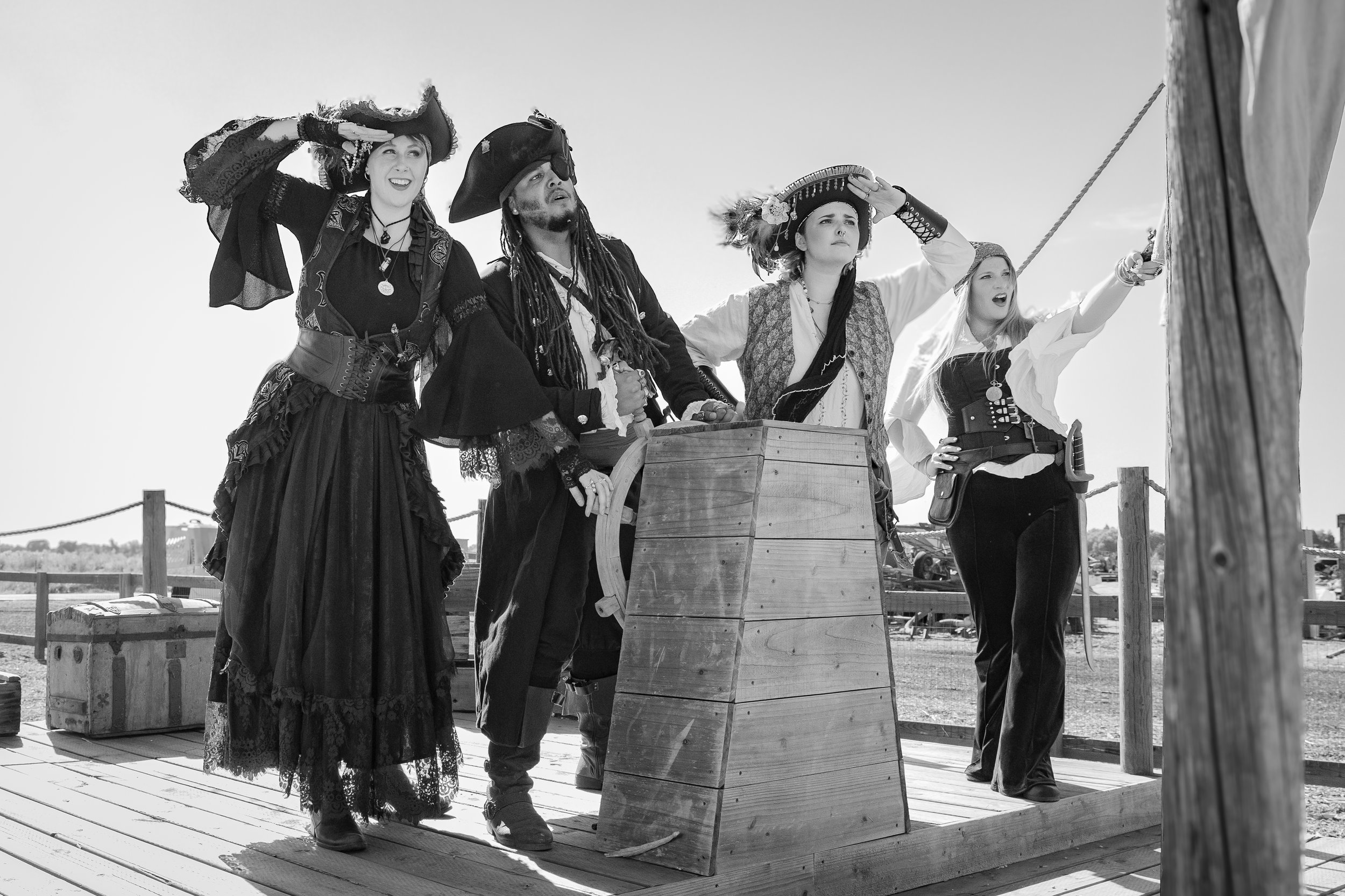 Actors dressed as pirates and a wench in pirate costumes on a ship deck, with one pointing a sword and another shielding her eyes, in a themed historical reenactment.
