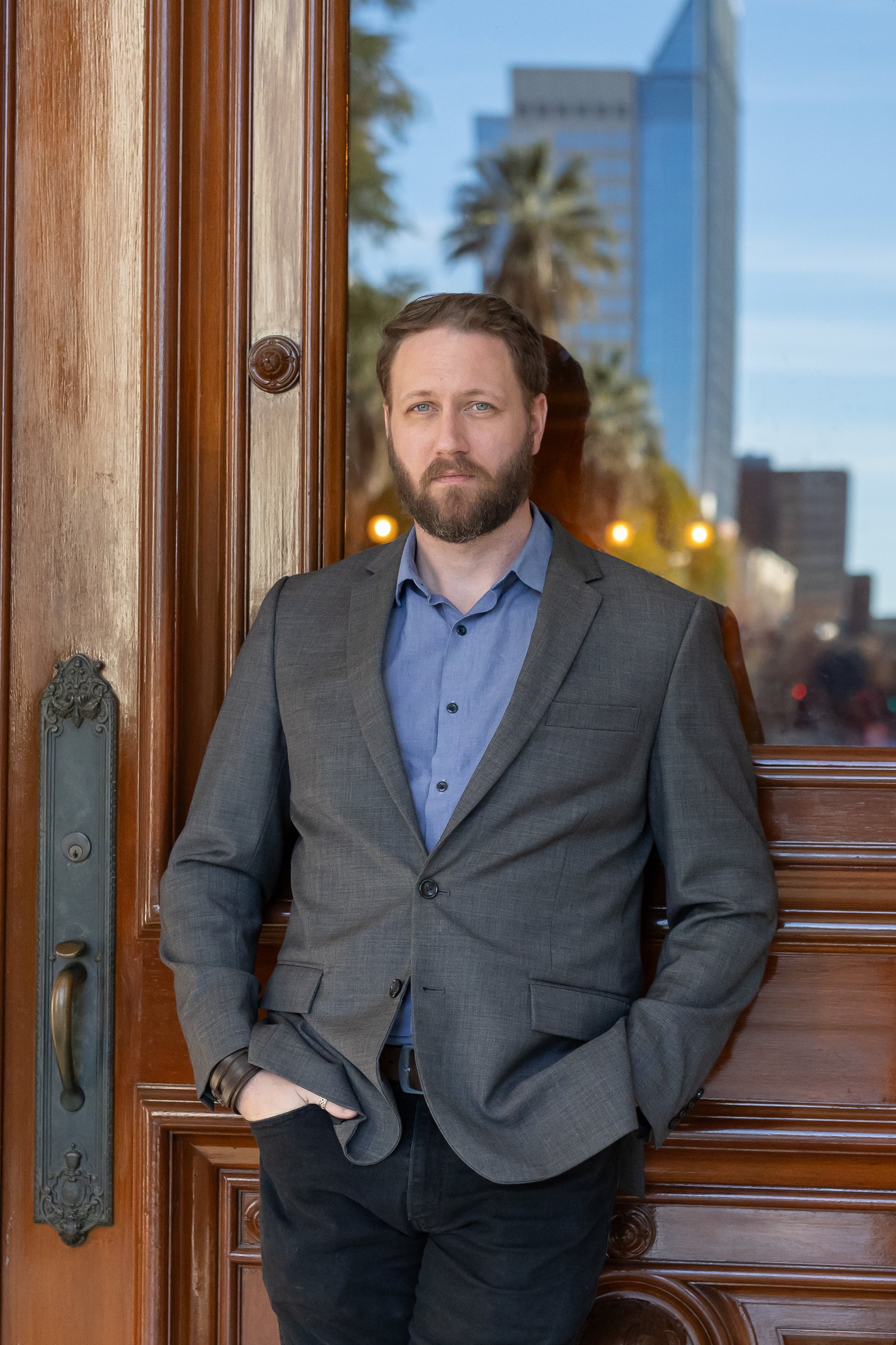 A man with light brown hair and a beard, dressed in a gray blazer and blue shirt, leaning against a wooden doorframe outside, with city buildings and palm trees visible in the background during dusk.