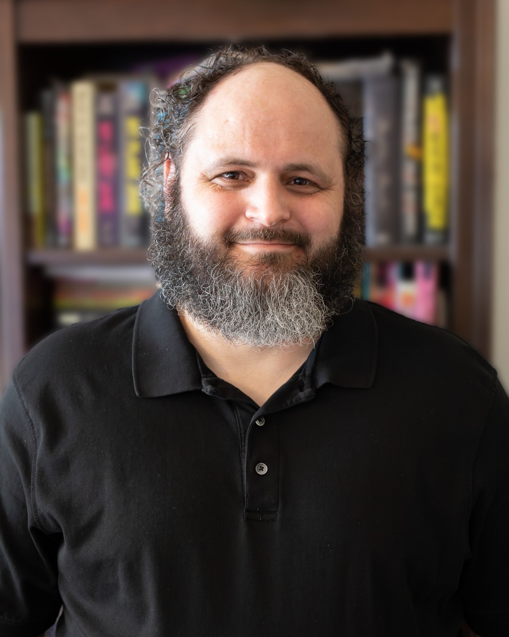 A man with a beard smiling, wearing a black polo shirt, standing in front of a bookshelf filled with colorful books.