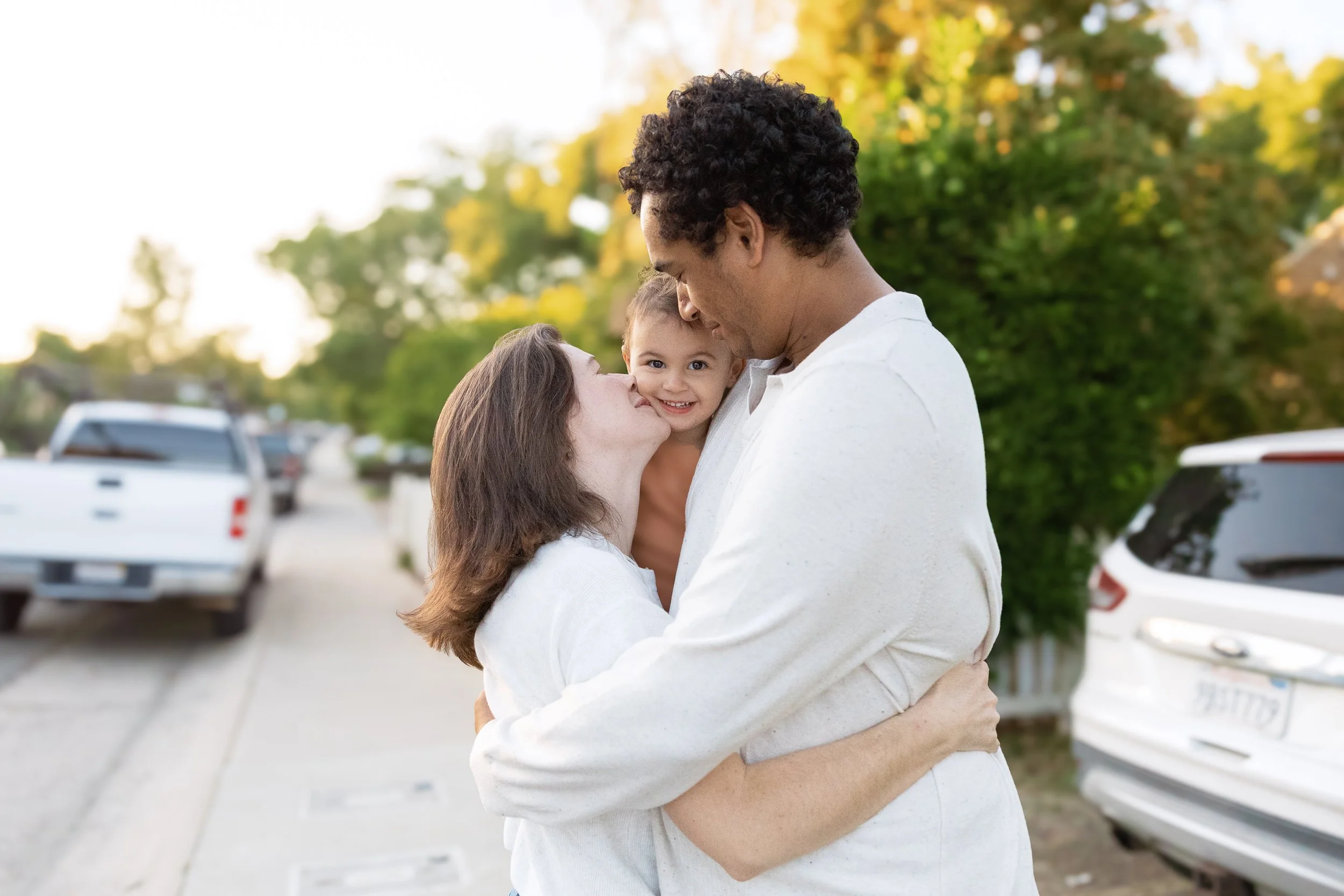 A family of three embracing outdoors during daytime, with trees and parked cars in the background. The woman and man are holding the young child, who is smiling at the camera.