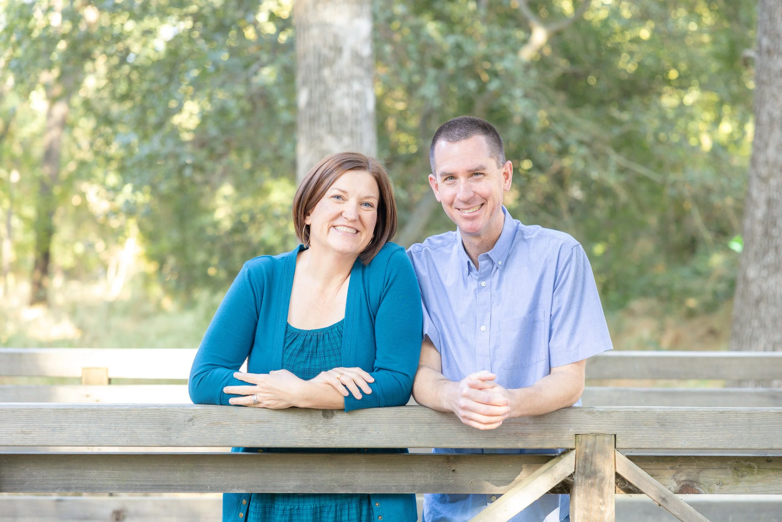 A man and woman smile while leaning on a wooden fence in a bright outdoor setting with trees and soft natural light behind them.