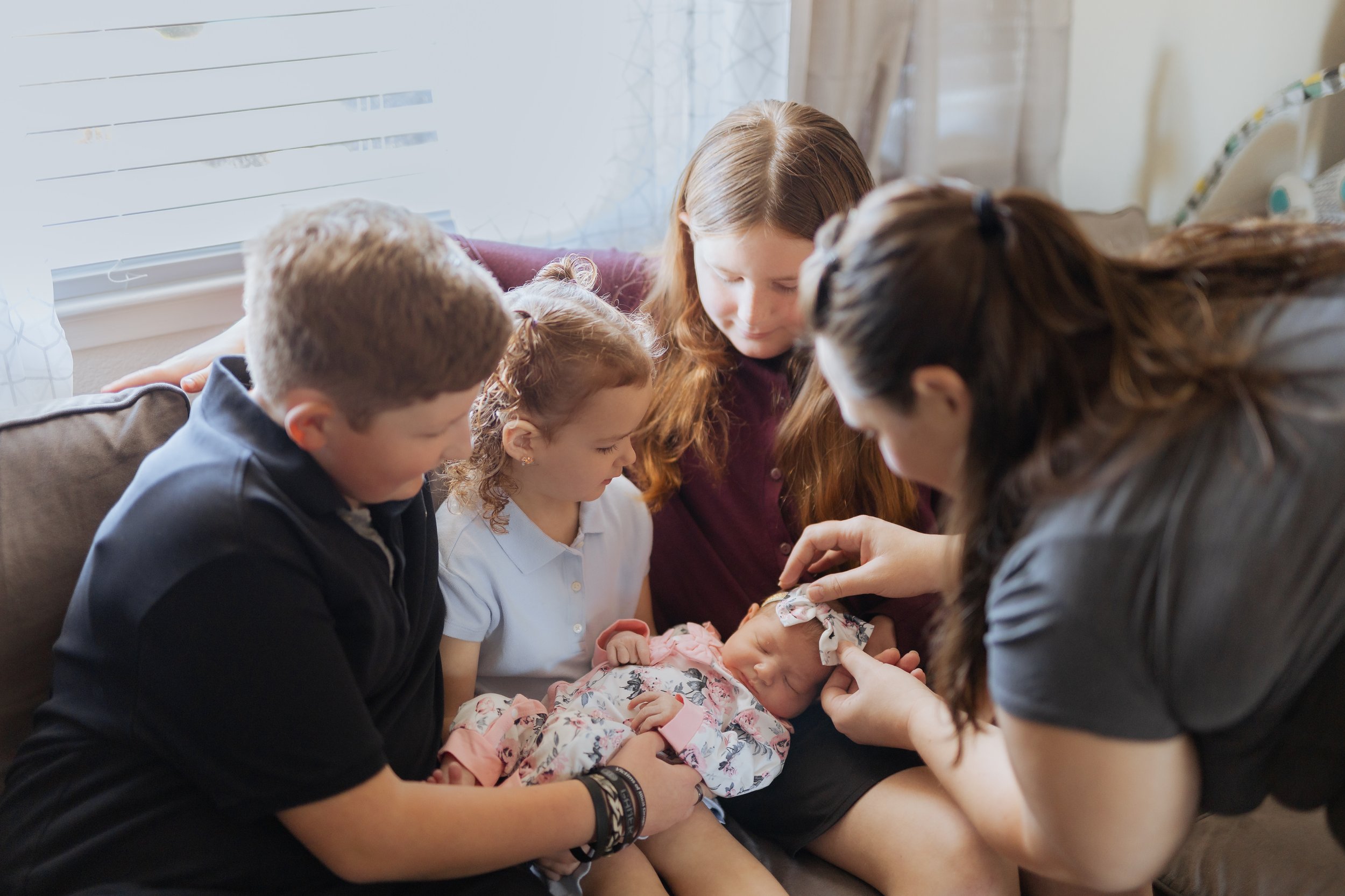 A group of five people, including four children and one woman, sitting on a couch together, with a child holding and tending to a newborn baby, in a cozy living room with light coming through a window.