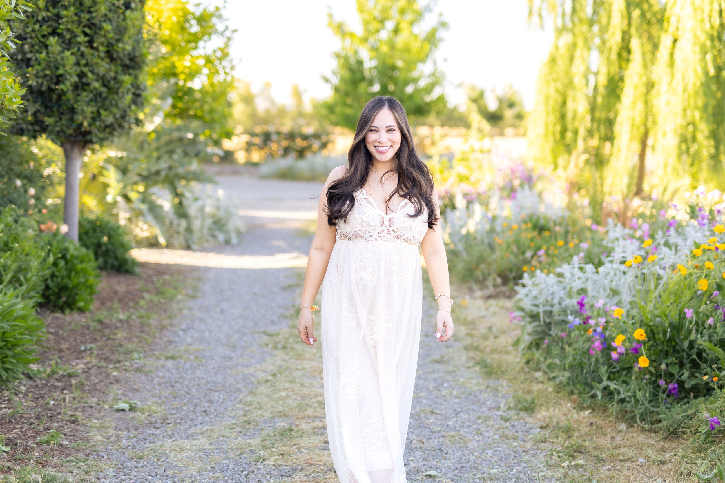 A woman in a white lace dress walking on a gravel path, smiling, surrounded by colorful flowers and lush green trees in a garden.