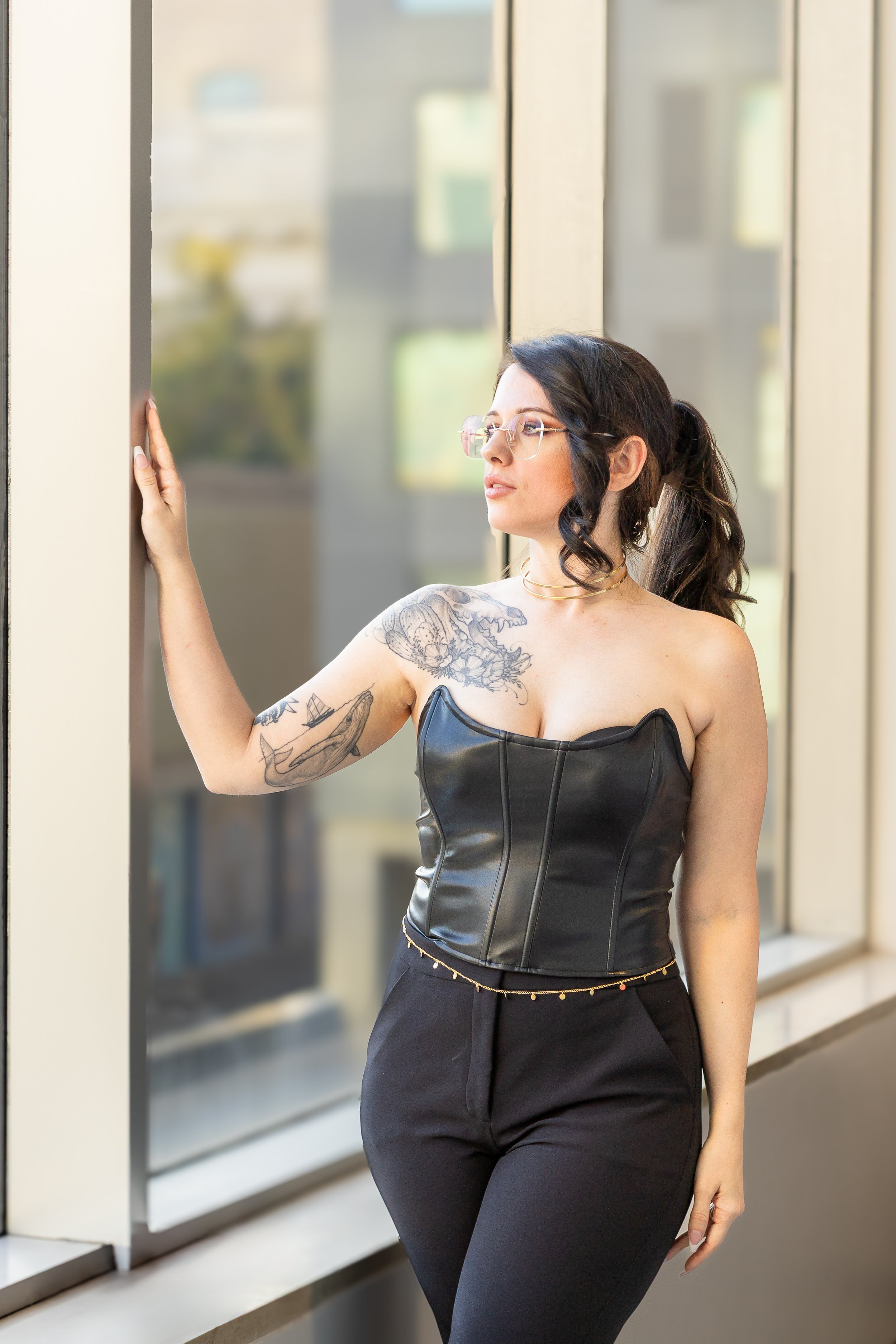 A woman with glasses, black hair in a ponytail, tattoos on her left arm, wearing a strapless black leather top and black pants, standing indoors by a window, touching the wall with her left hand and looking outside.