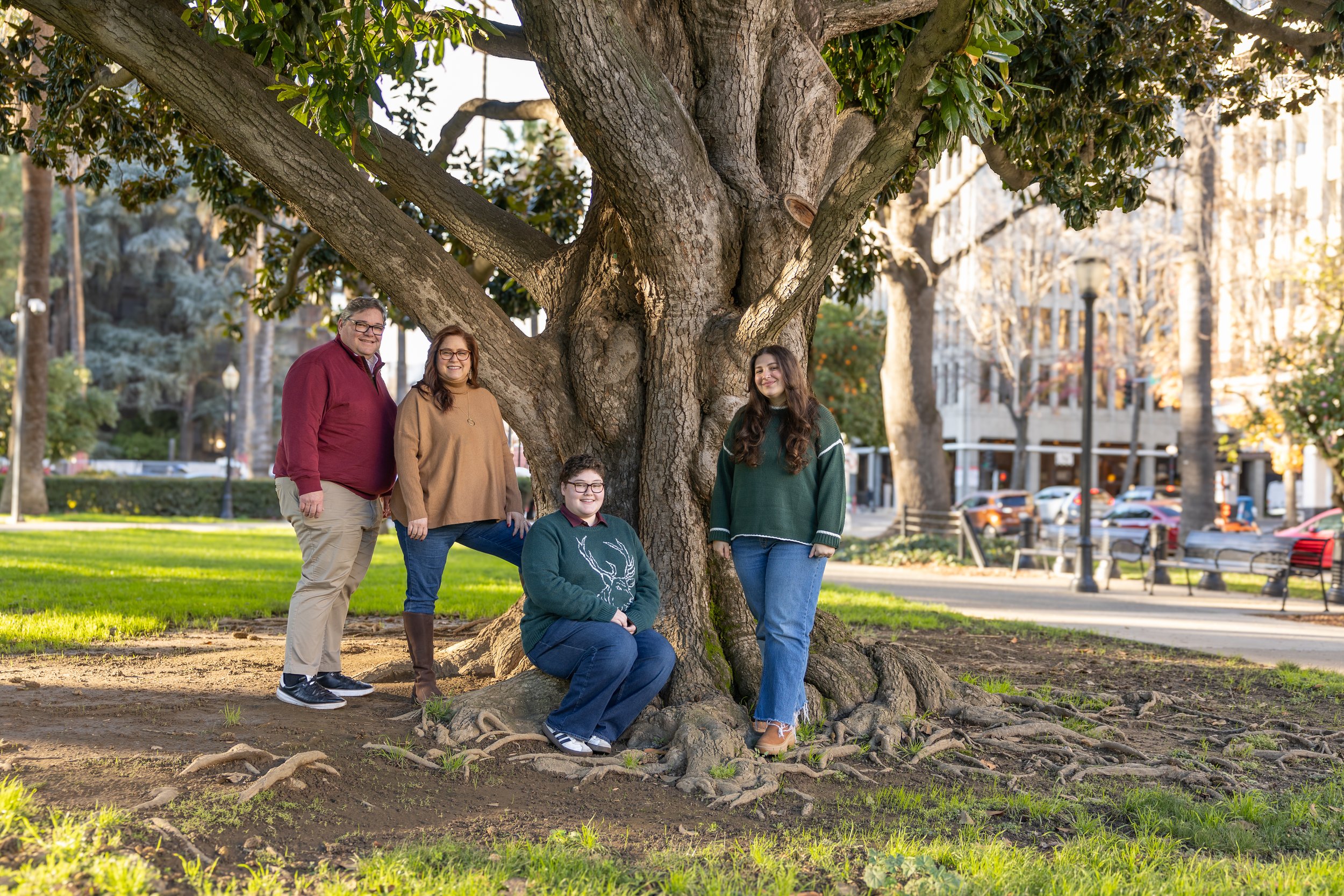 Family of four posing around a large tree in a park during daytime, with buildings and street in the background.
