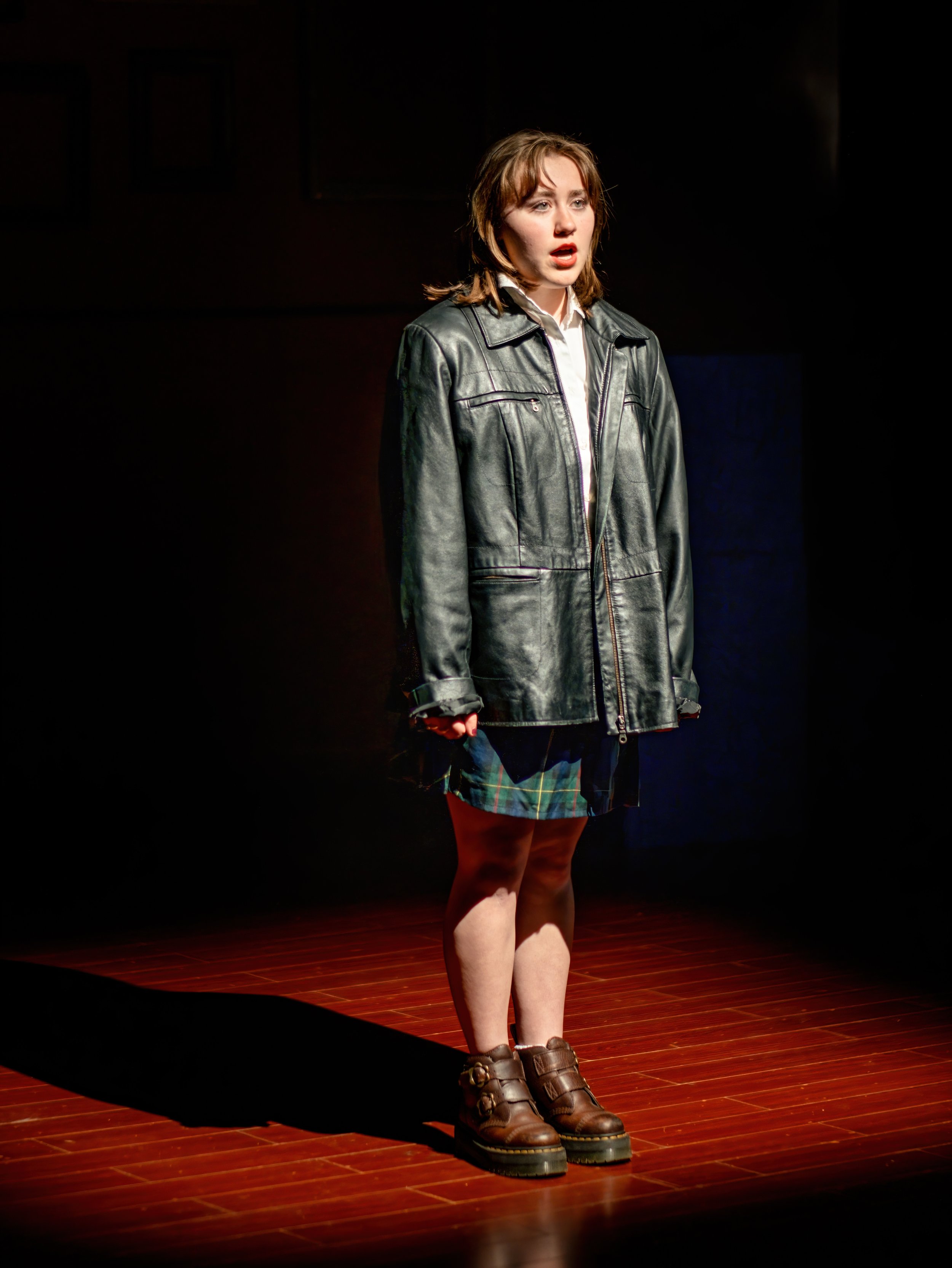 Young woman standing on stage in spotlight, wearing a black leather jacket, white shirt, plaid skirt, and brown boots, with a concerned or emotional expression.