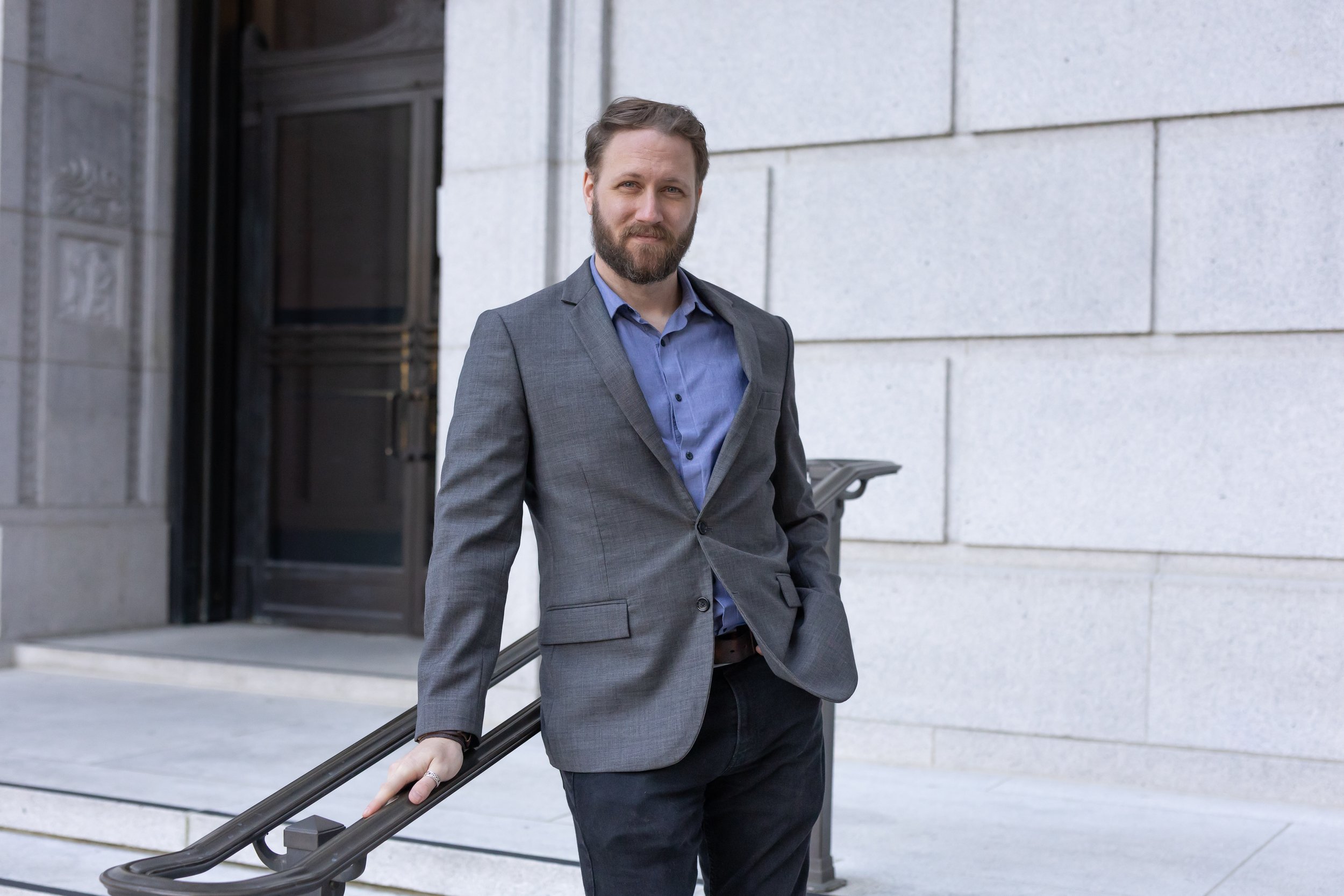 A man in a gray suit jacket and blue shirt standing outside a stone building, leaning on a black railing.