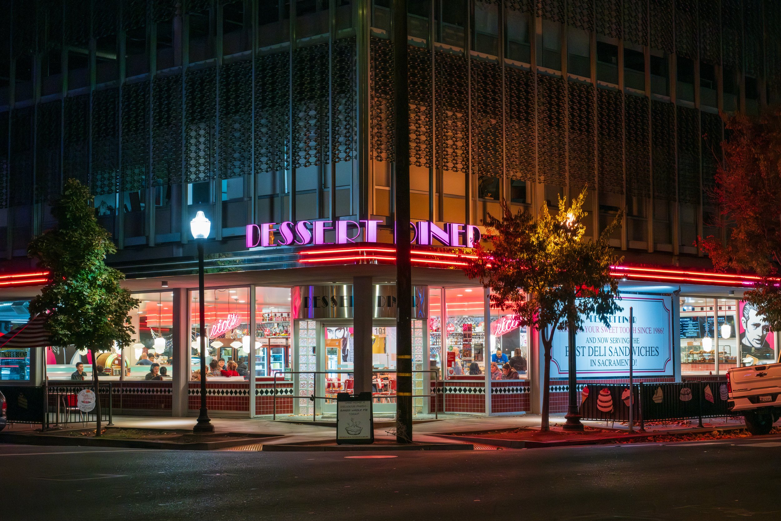 Night view of a brightly lit dessert shop with pink and purple neon signs, large window displaying people dining inside, and a digital sign with an advertisement outside.