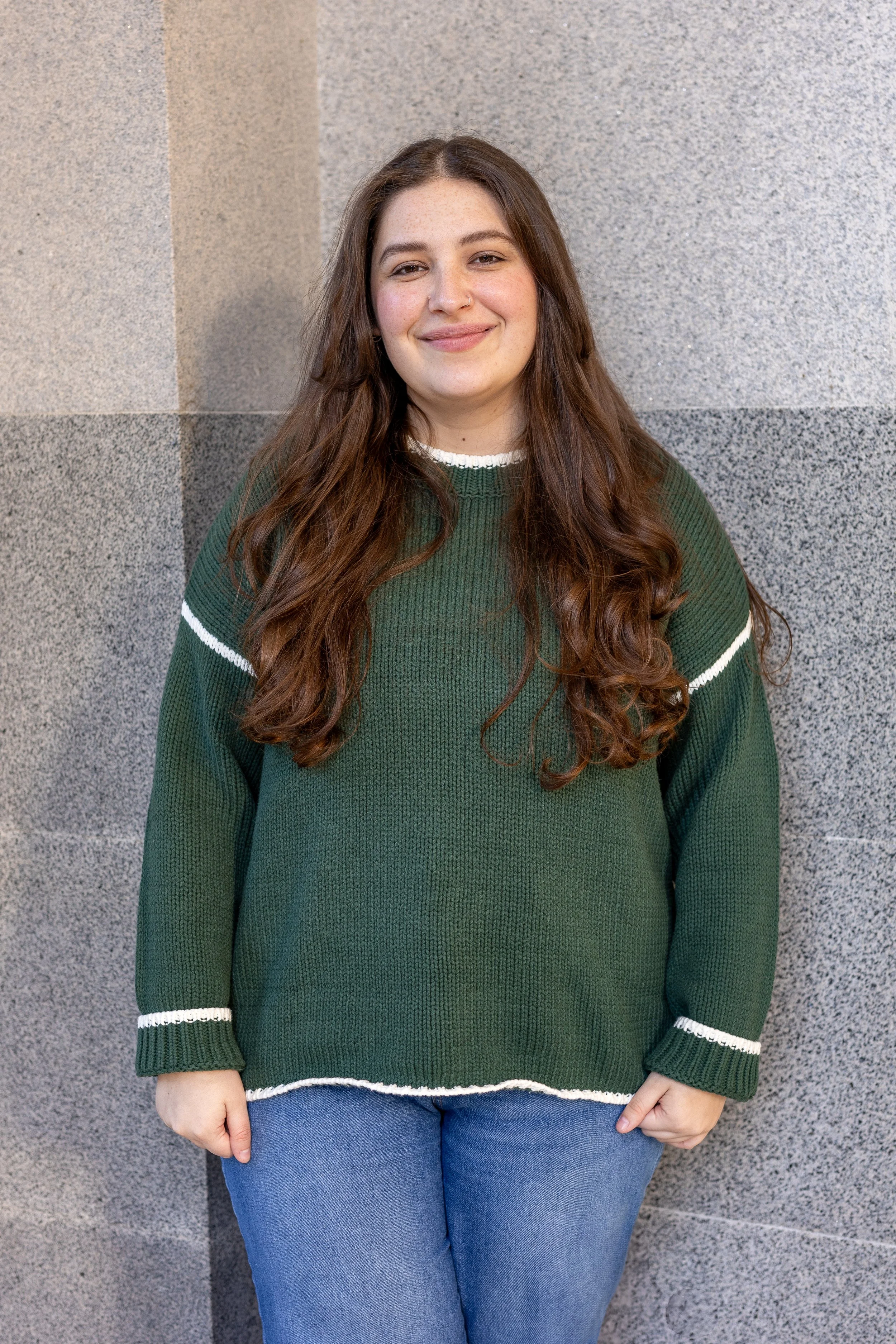 A young woman with long wavy brown hair, wearing a green sweater with white stripes at the neckline, cuffs, and hem, and blue jeans, standing against a textured stone wall background.