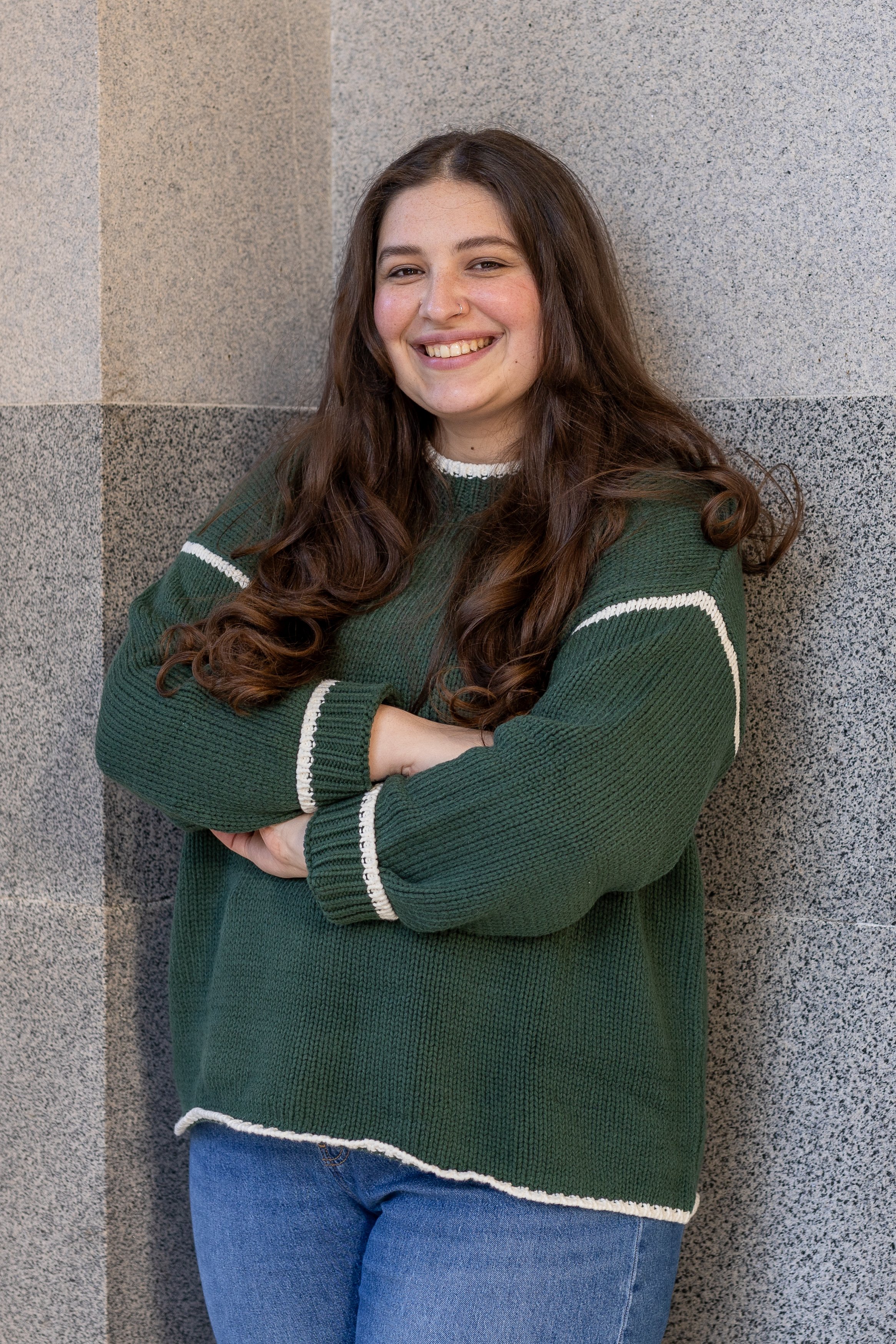 A young woman with long, curly brown hair, smiling and standing with arms crossed, wearing a green sweater with white accents and blue jeans, leaning against a tiled wall.