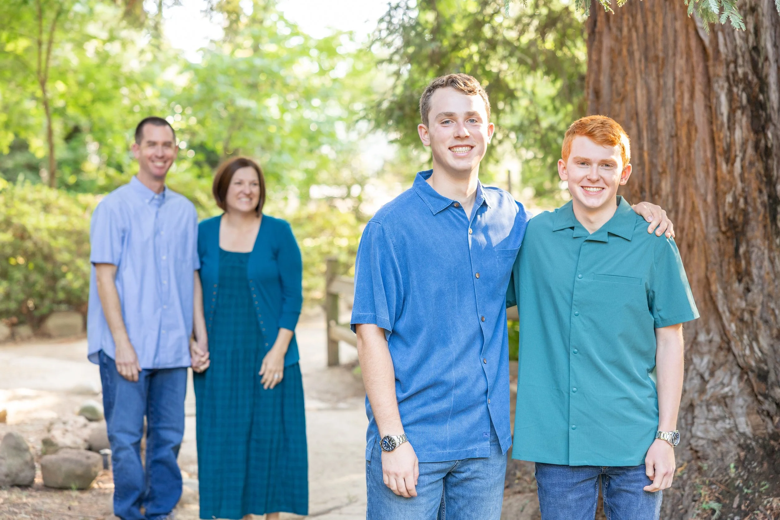 A family of four walks side-by-side on a forest path, with the parents holding hands in the background and two teenage sons smiling at the camera in the foreground.