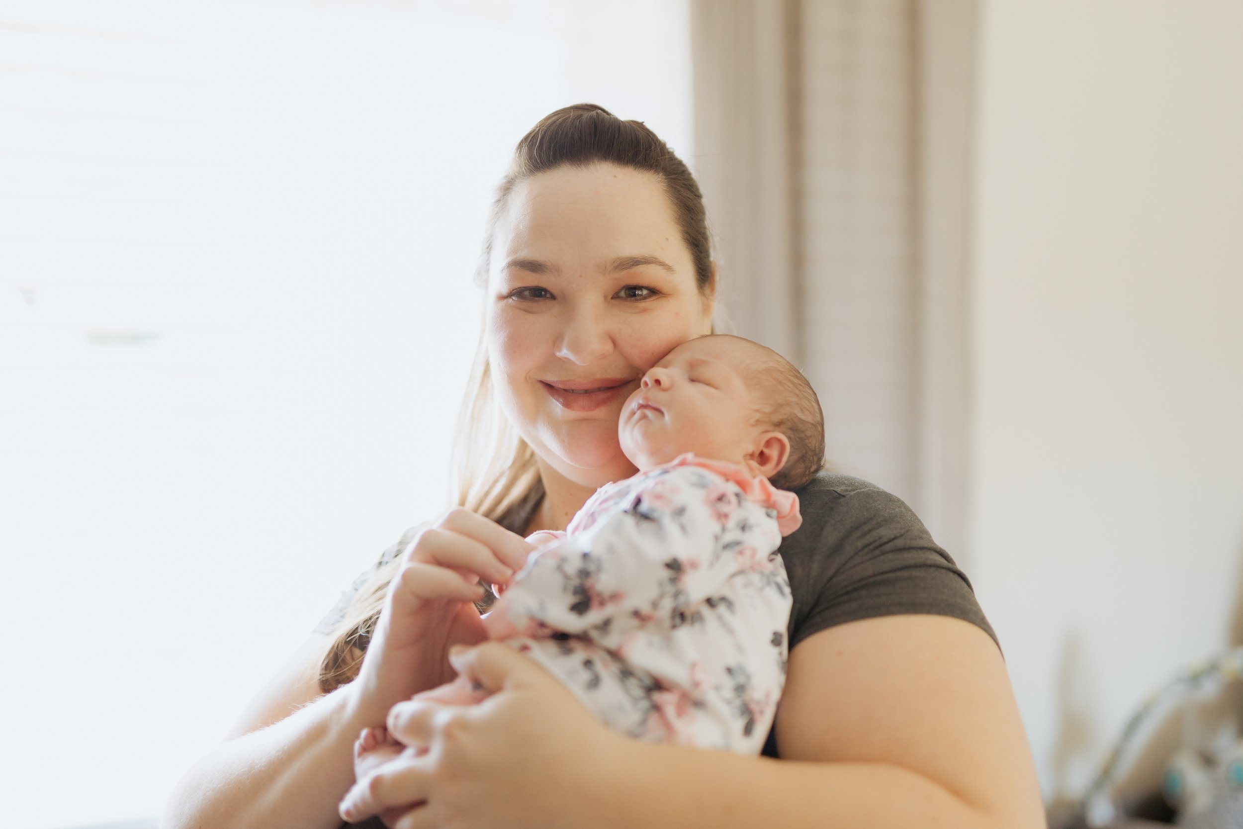 A woman smiling and holding a sleeping baby close to her cheek indoors.