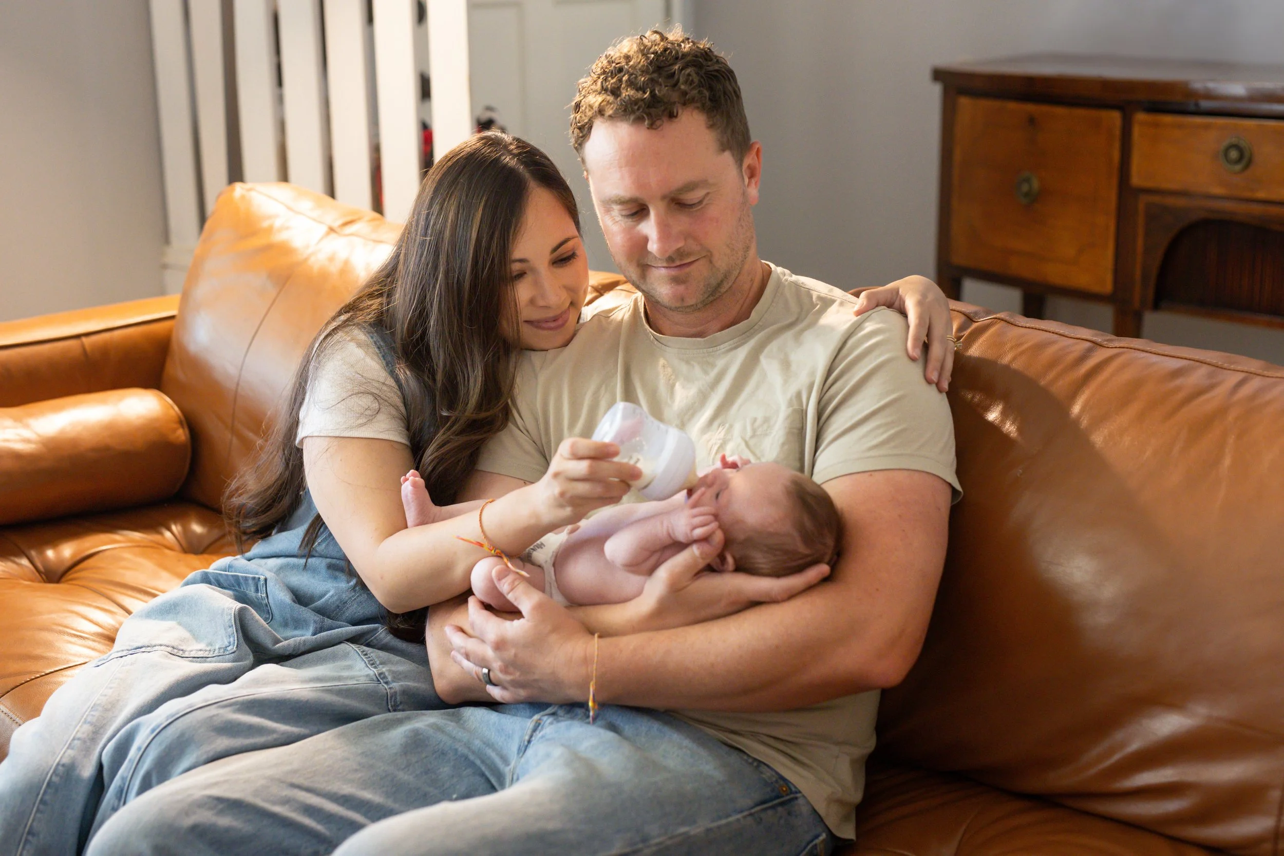 A couple sitting on a leather couch, feeding a baby, with the woman smiling and leaning into the man, who is holding the baby.