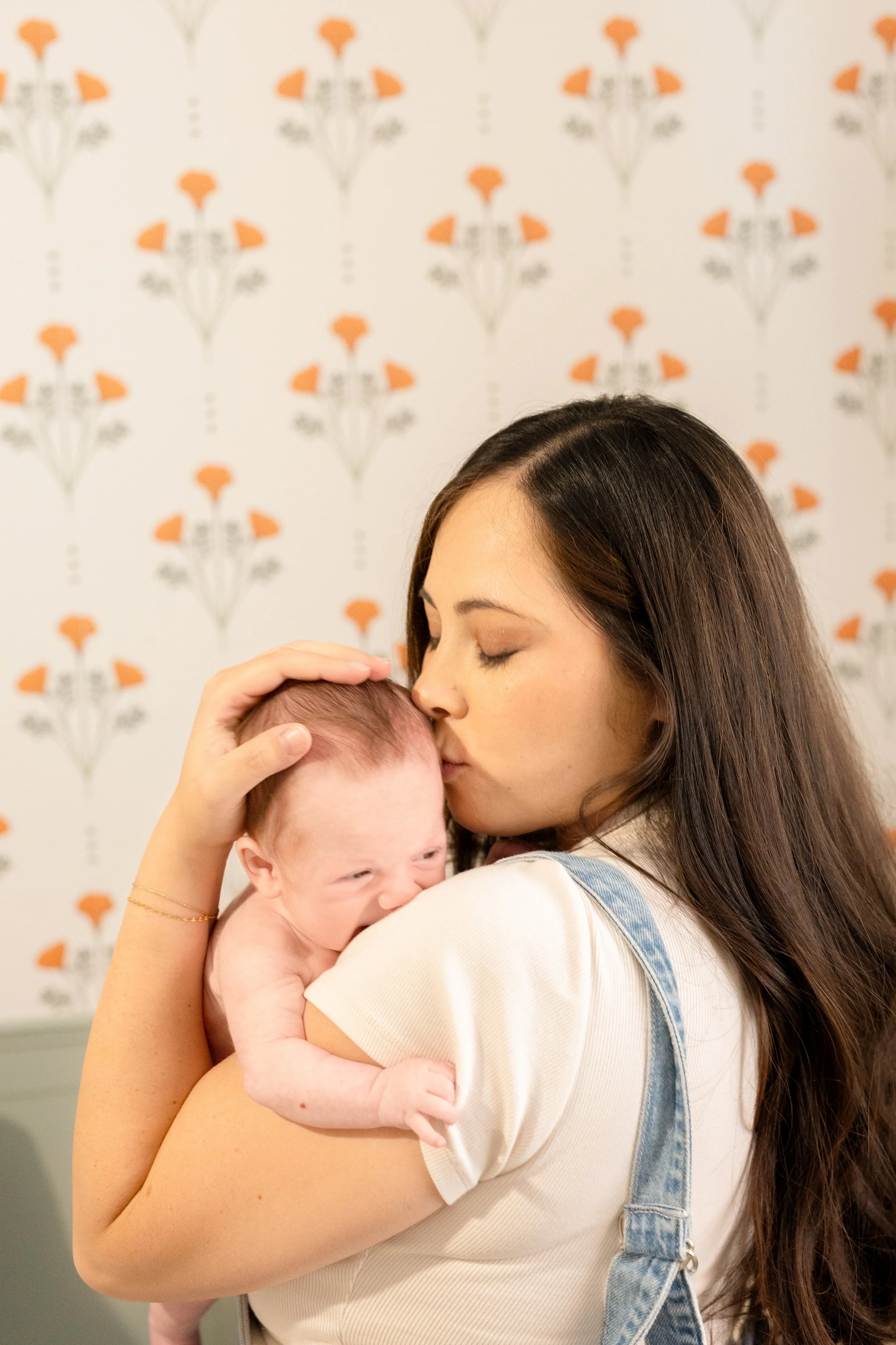 A woman with long brown hair holding a laughing baby with light skin, set against a background with a floral pattern.