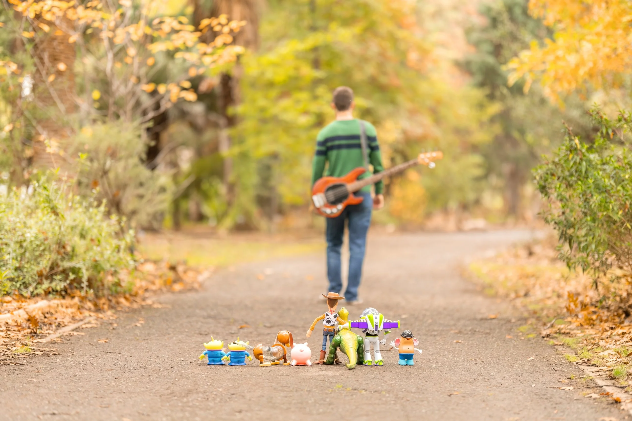 Toy figurines of animals and characters placed on a tree-lined path in fall. In the background, a person with a guitar walks away, surrounded by autumn-colored trees.