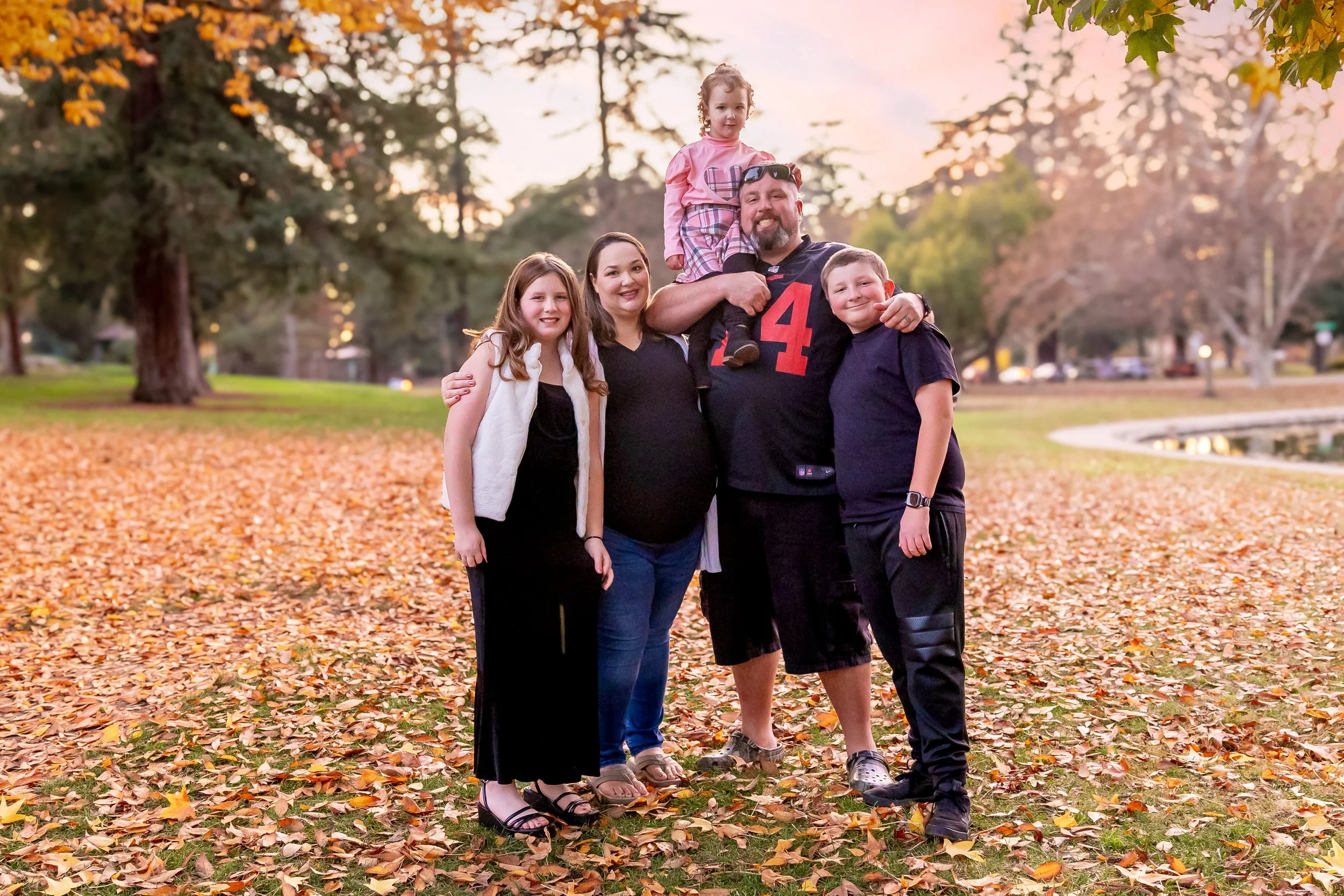 Family of five posing outdoors in a park with autumn leaves on the ground, during sunset.