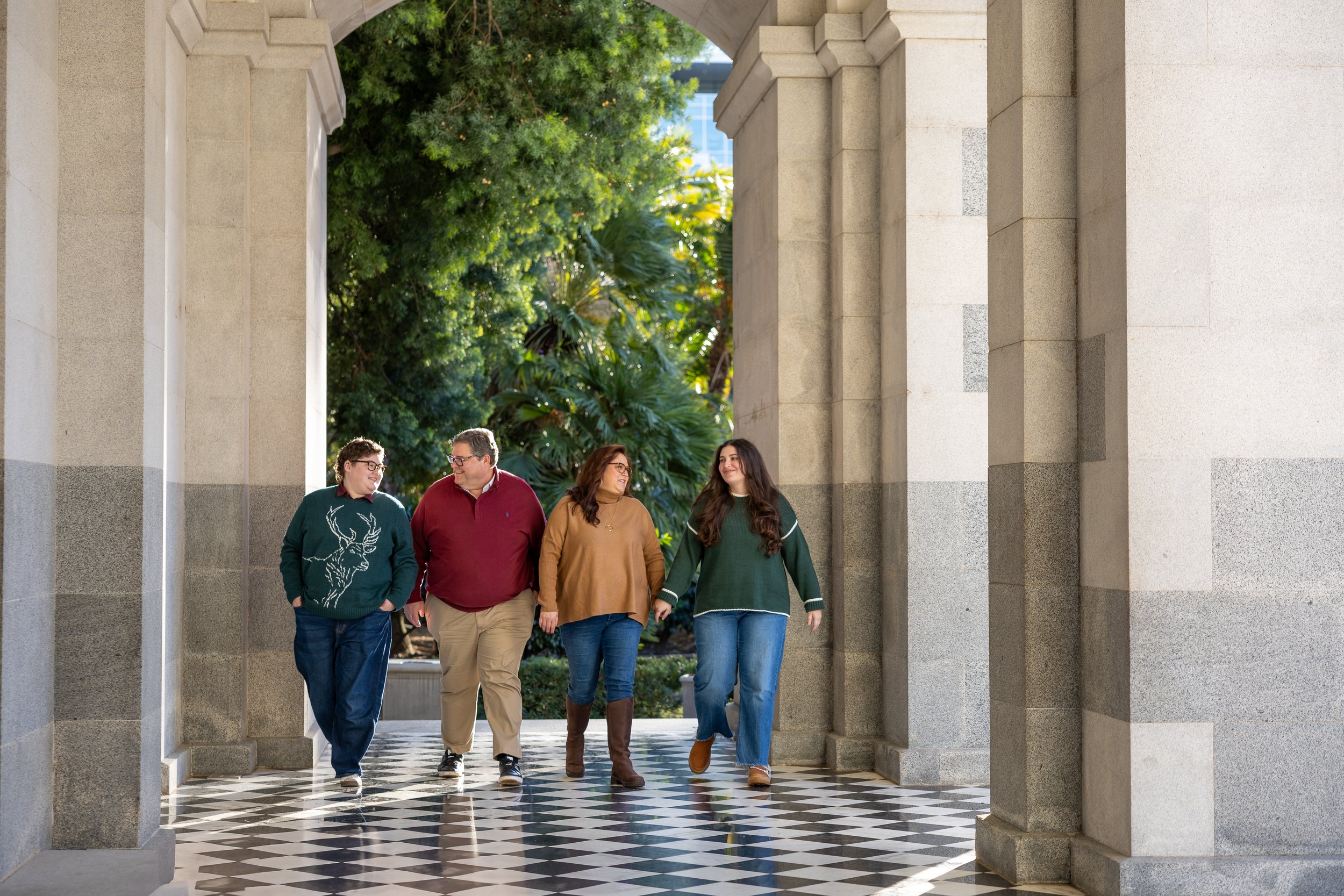 Four people walking together under an arched doorway with lush green trees in the background. They are smiling and engaged in conversation, dressed casually.