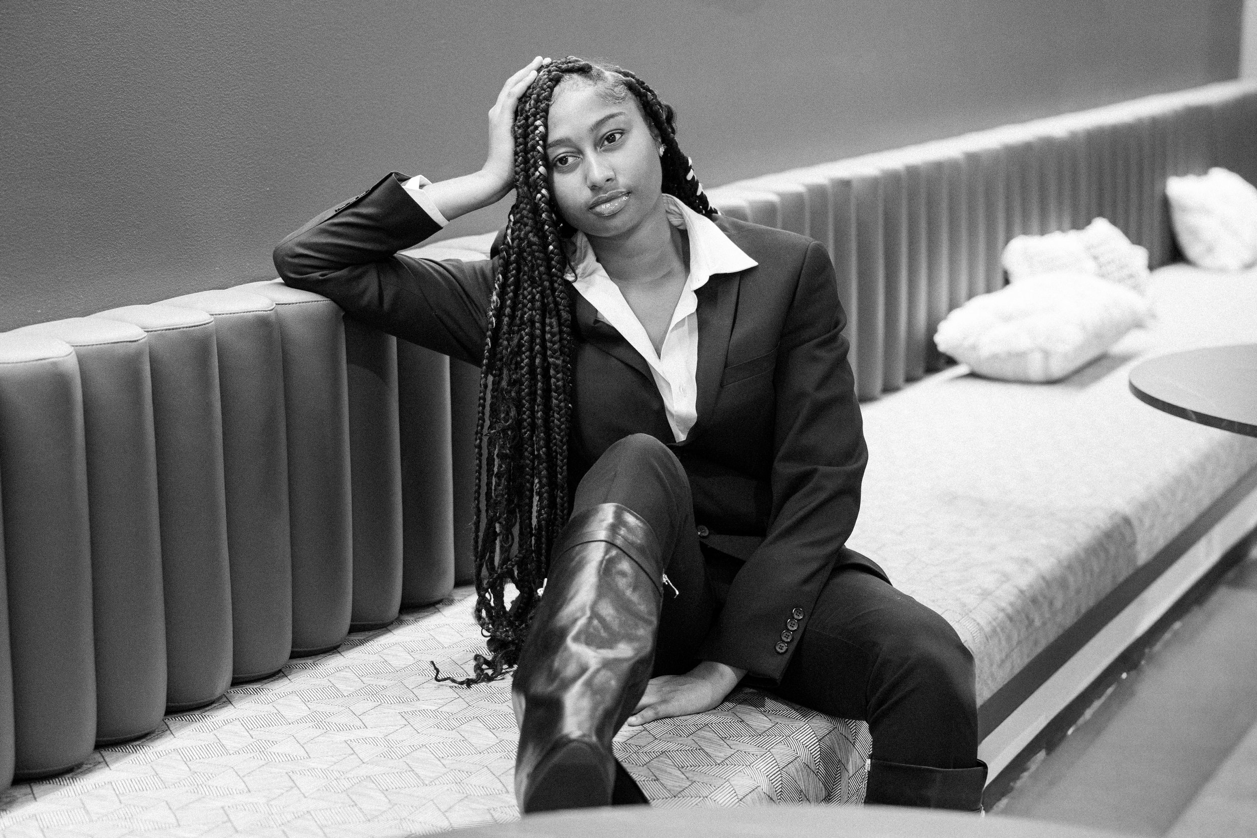 A woman dressed in a suit, sitting on a bench in a lounge, with one leg crossed over the other, her hand resting on her head, looking thoughtful.