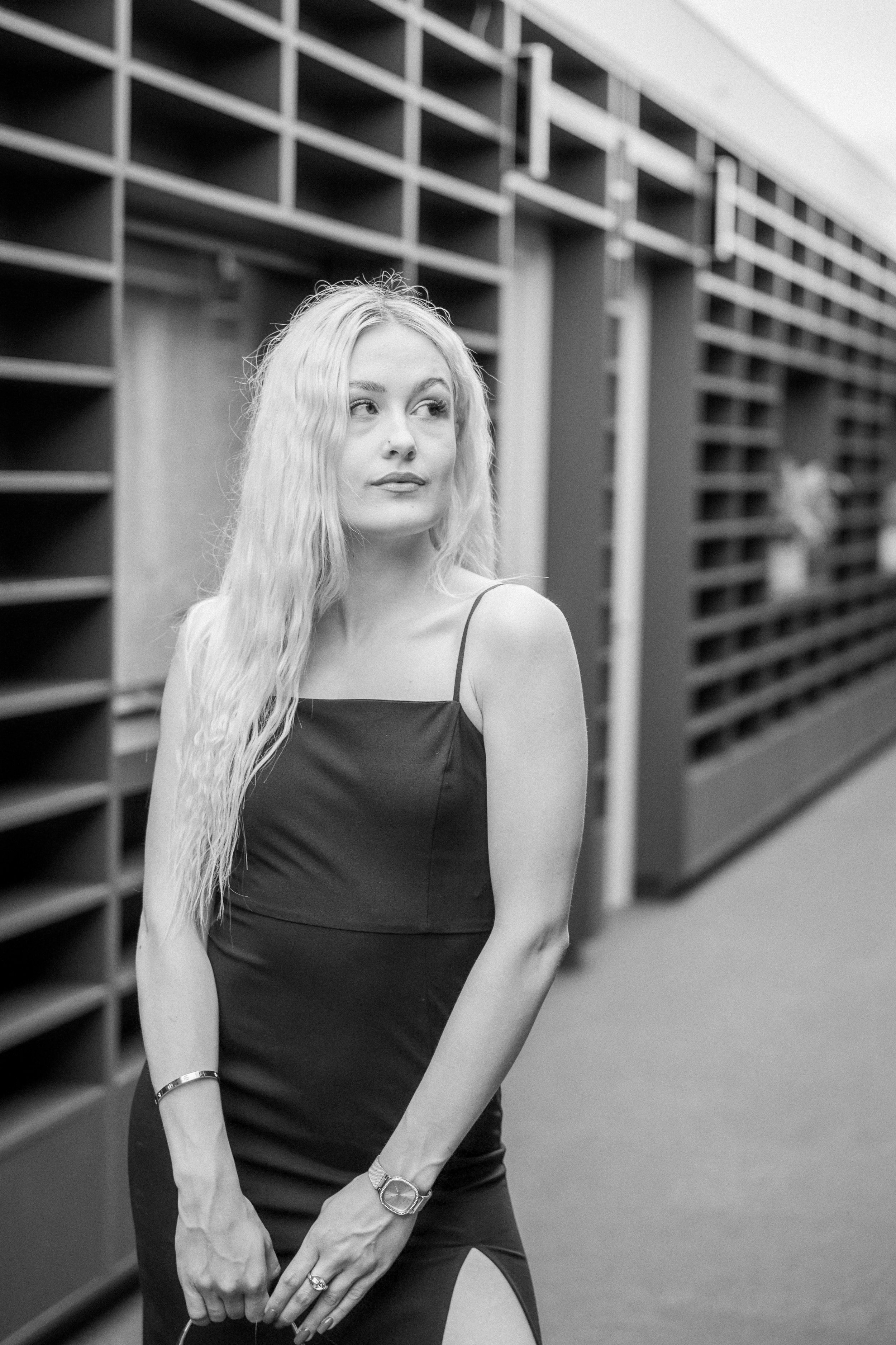 A young woman with long, wavy blonde hair wearing a black dress with spaghetti straps, standing in a hallway with multiple empty wooden mailboxes or cubbies on the walls.