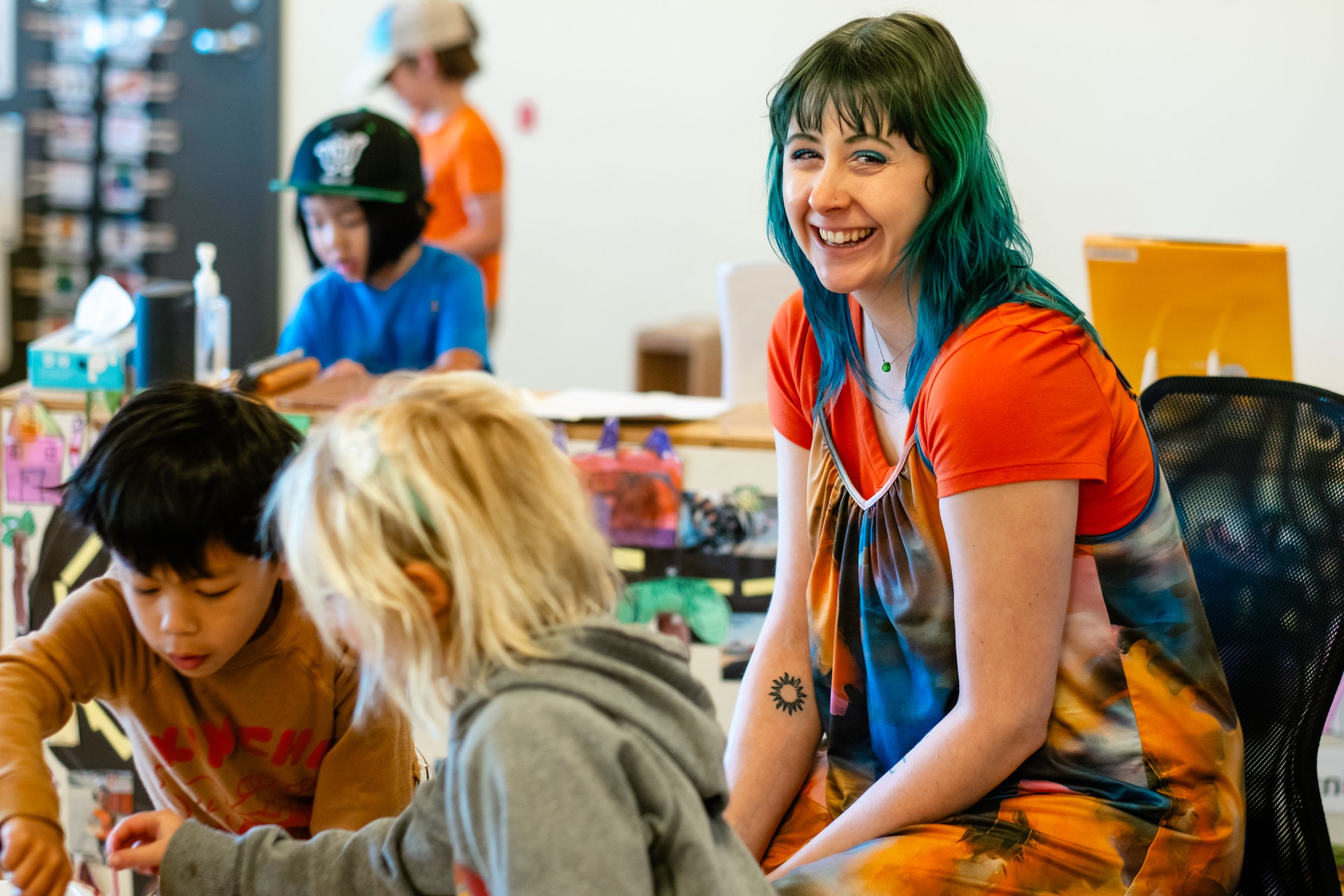 A smiling woman with blue and green hair, wearing an orange and multicolored dress, sitting and interacting with children in a classroom setting.