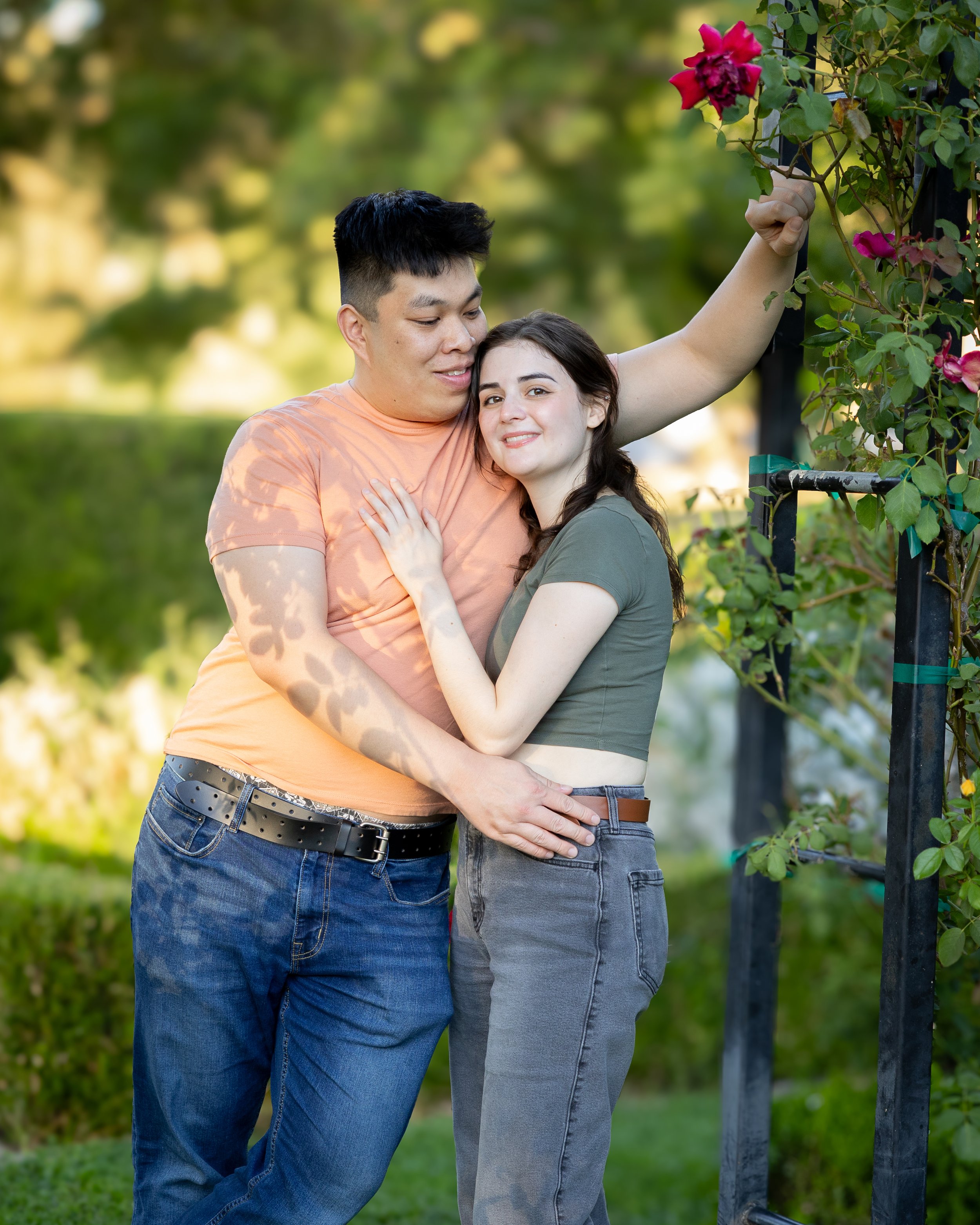 A young couple hugging outdoors next to a pink rose bush, with blurred greenery in the background.
