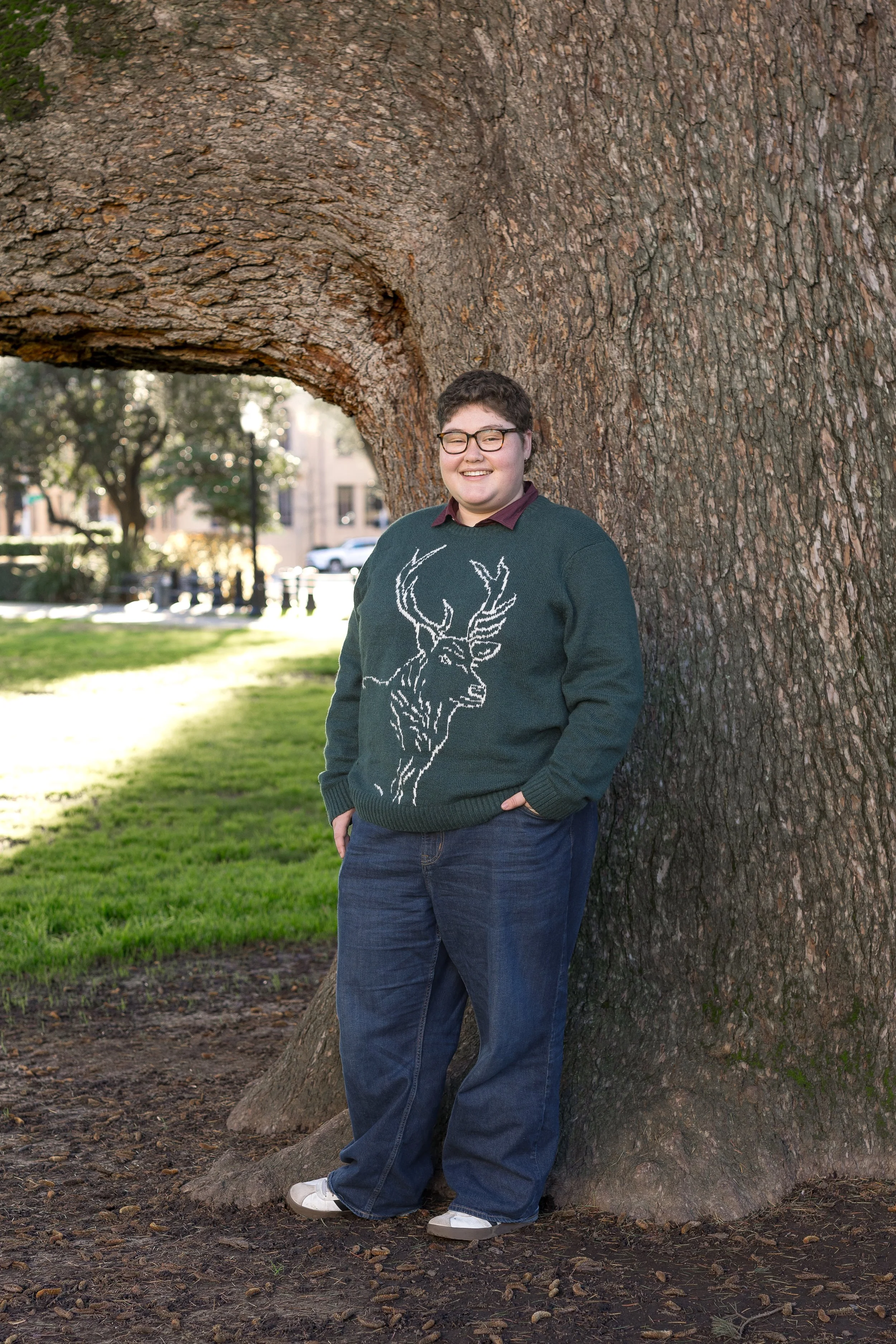 A person wearing glasses and a green sweater with a white deer design, standing outdoors near the base of a large tree with textured bark. The person has short, dark hair and is smiling, with a park and city buildings in the background.