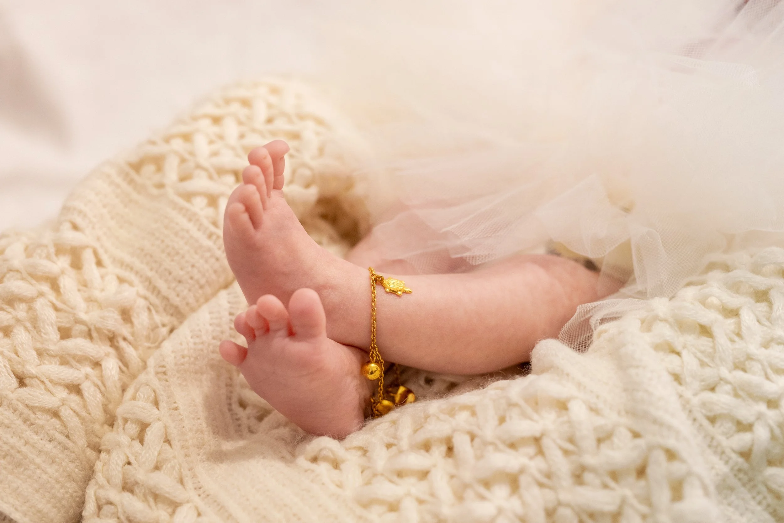 Close-up of a baby's feet with a gold bracelet, resting on a soft, cream-colored knitted blanket.
