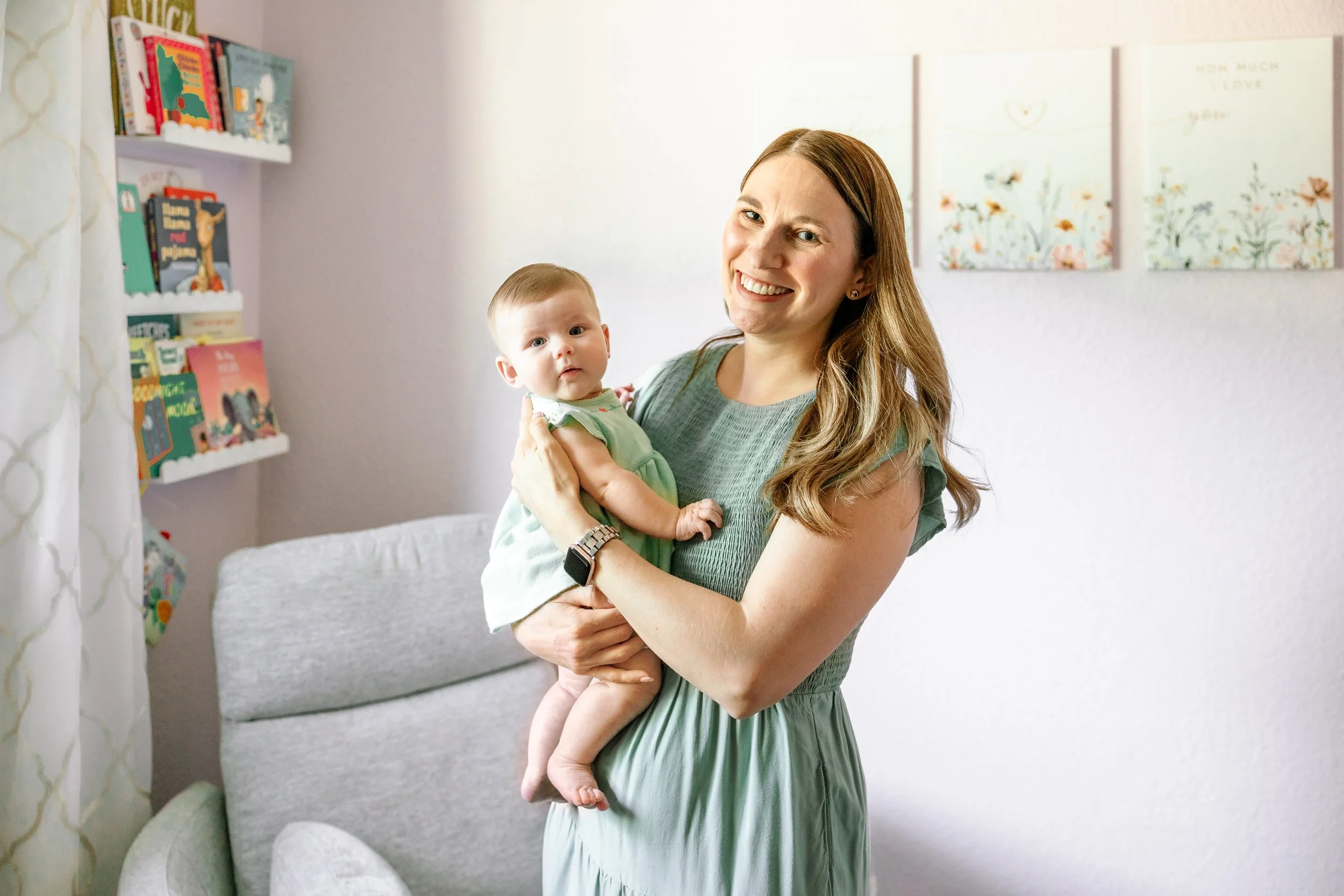 A woman with long, light brown hair smiling while holding a baby girl with light skin and blue eyes in a cozy, decorated room. The woman is wearing a green dress, and the baby is wearing a light-colored dress. There are books on a shelf and floral wa