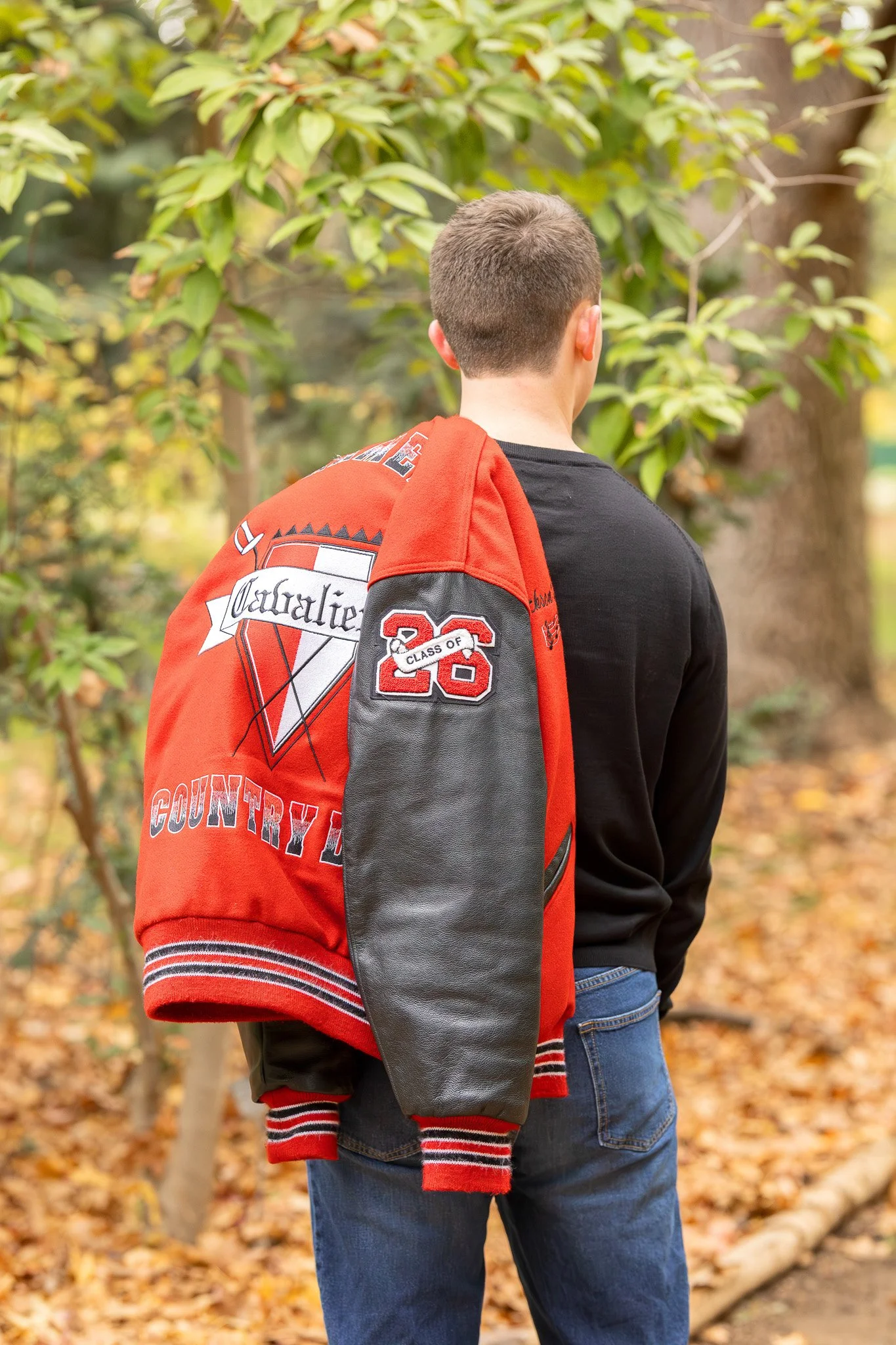 A person with short hair wearing a black shirt and blue jeans, with a red and black letterman jacket draped over their shoulder, standing outdoors among autumn trees with fallen leaves.