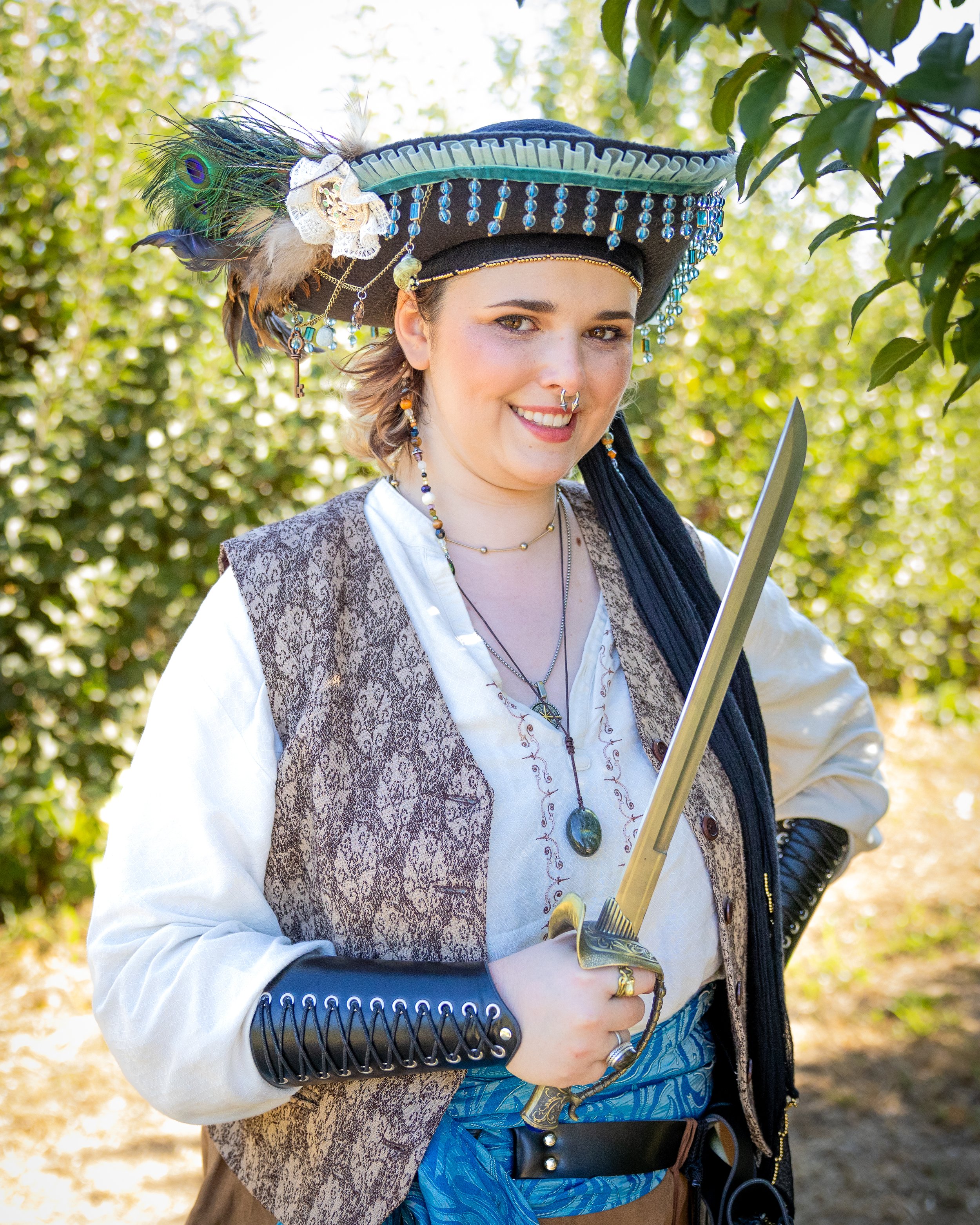 A woman dressed as a pirate, wearing a feathered hat with beads, jewelry, and holding a sword outdoors in a sunny, green environment.