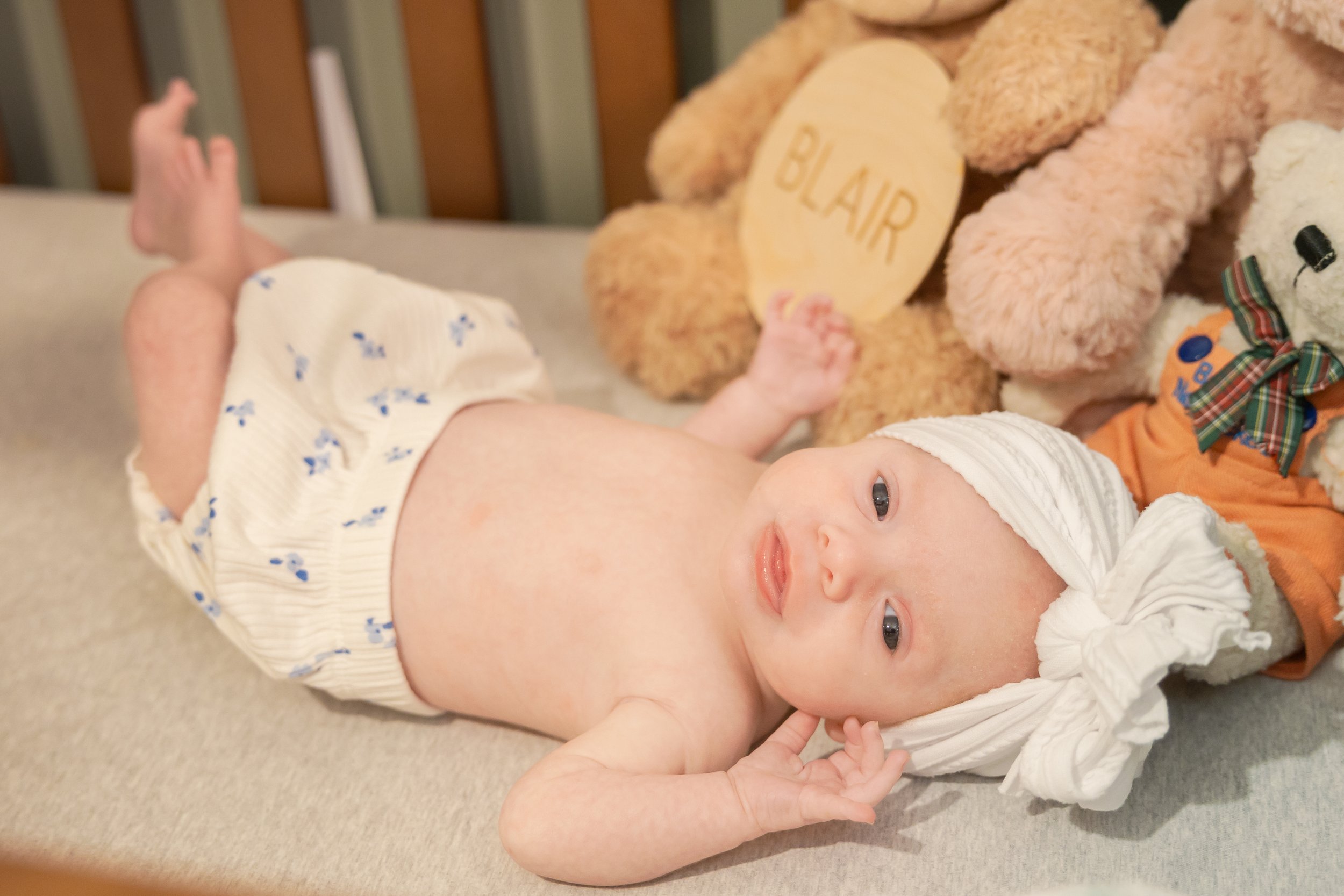A baby lying on its back on a beige carpet, wearing a white hat and a white outfit with small blue patterns, smiling and looking at the camera with wide eyes, surrounded by teddy bears.