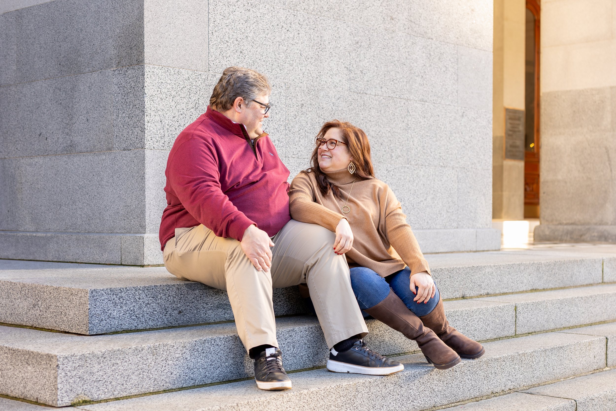 A man and woman sitting together on stone steps outside a building, engaged in conversation and smiling at each other.