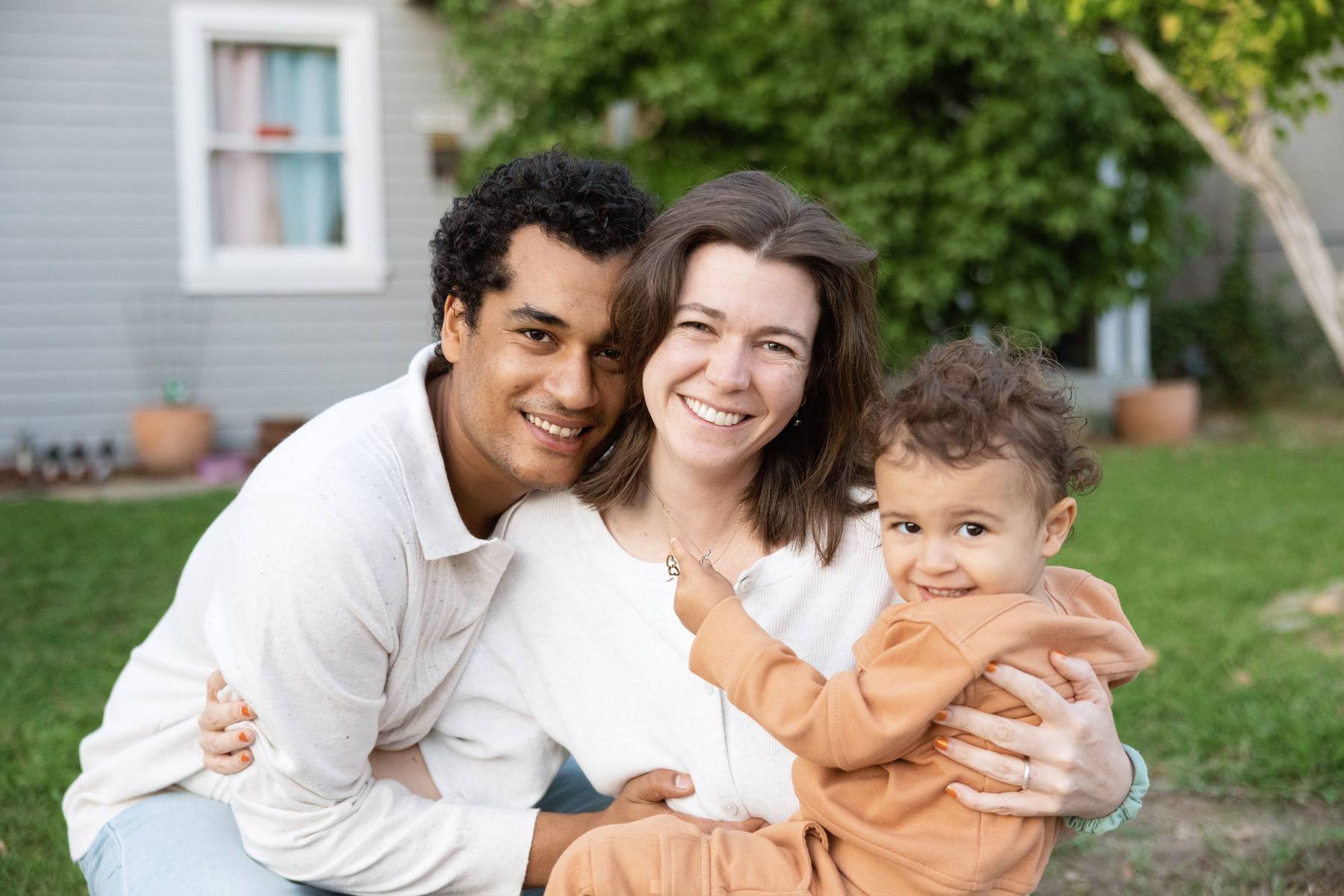 A mother and father sit closely together in a backyard, smiling while their toddler sits on their lap and looks at the camera.