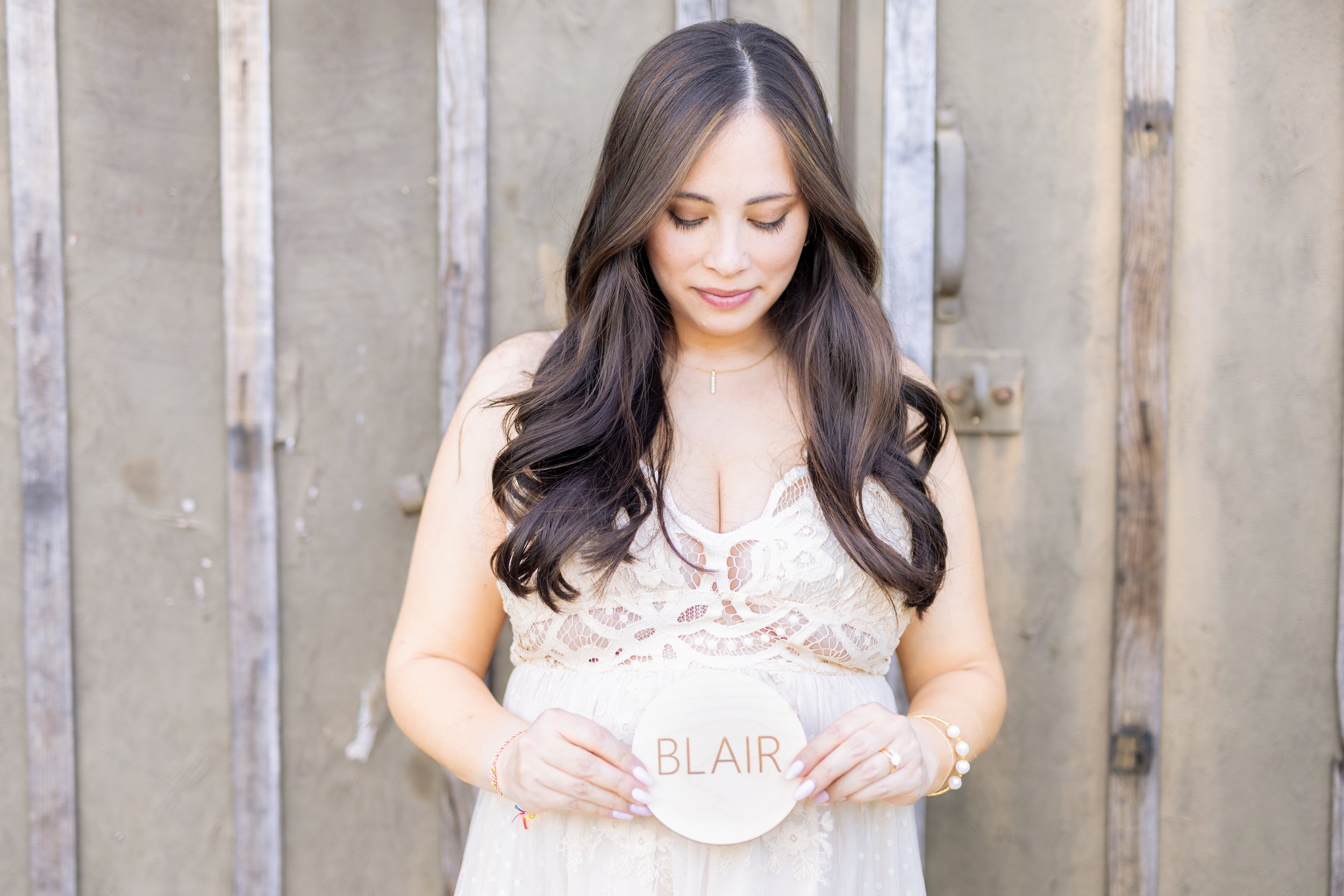 Woman with long dark hair in a cream dress standing in front of a wooden fence, holding a white round sign with the name 'BLAIR' in red letters.