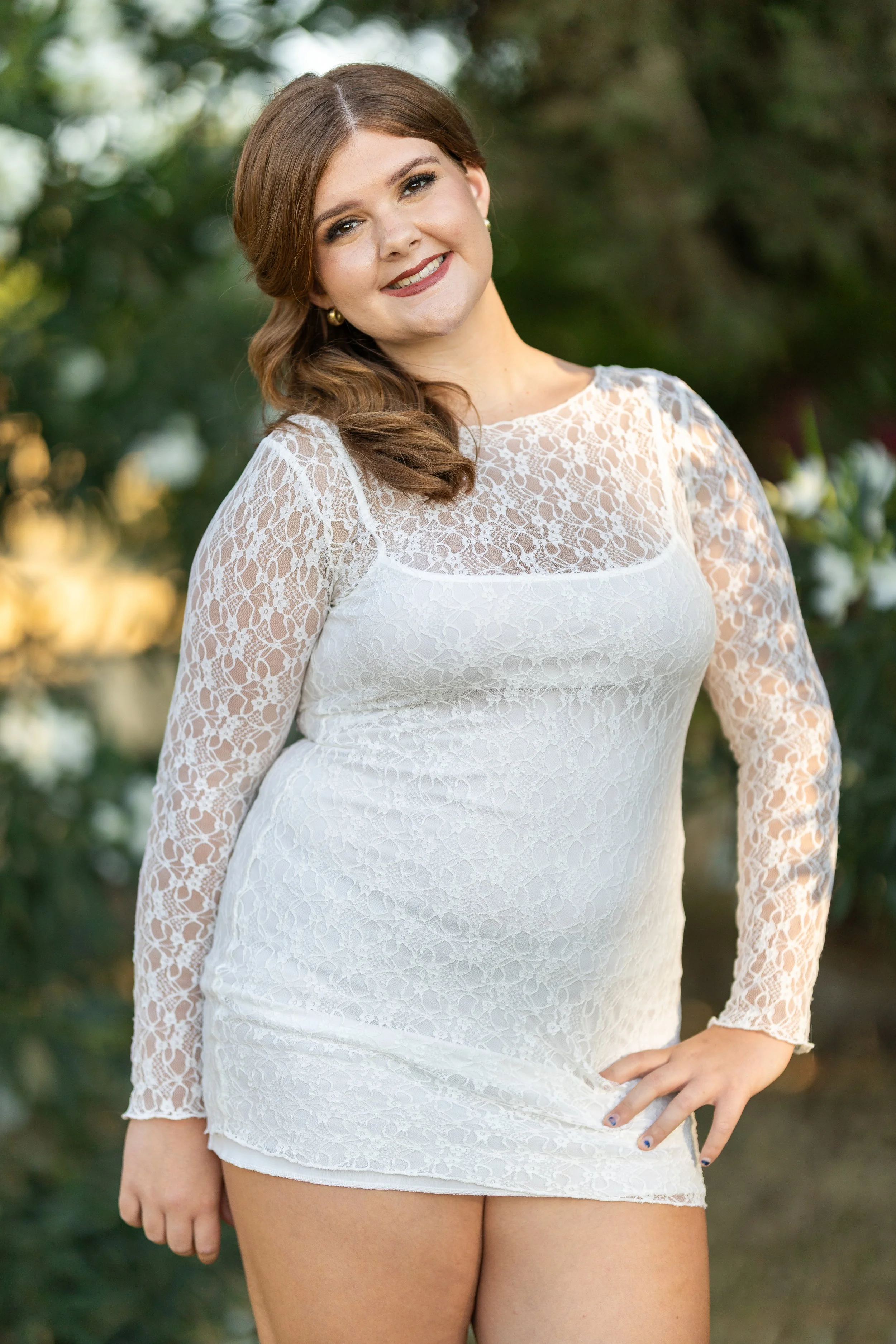 A woman wearing a white lace dress in an outdoor setting with greenery in the background.