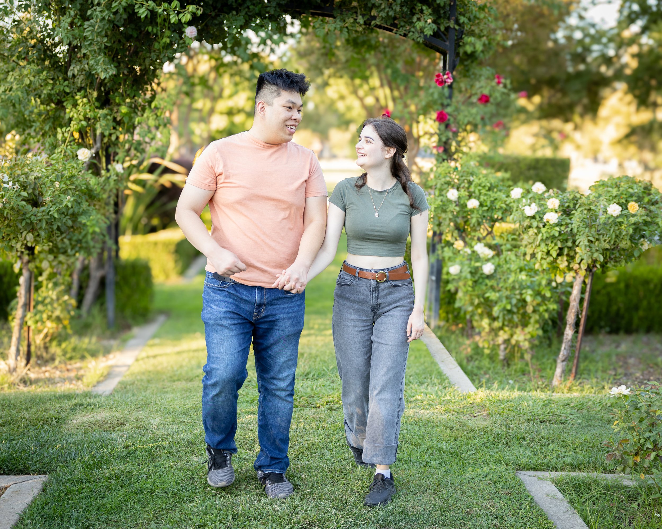 A young man and woman walking hand-in-hand in a garden with green foliage and blooming roses, smiling and looking at each other.