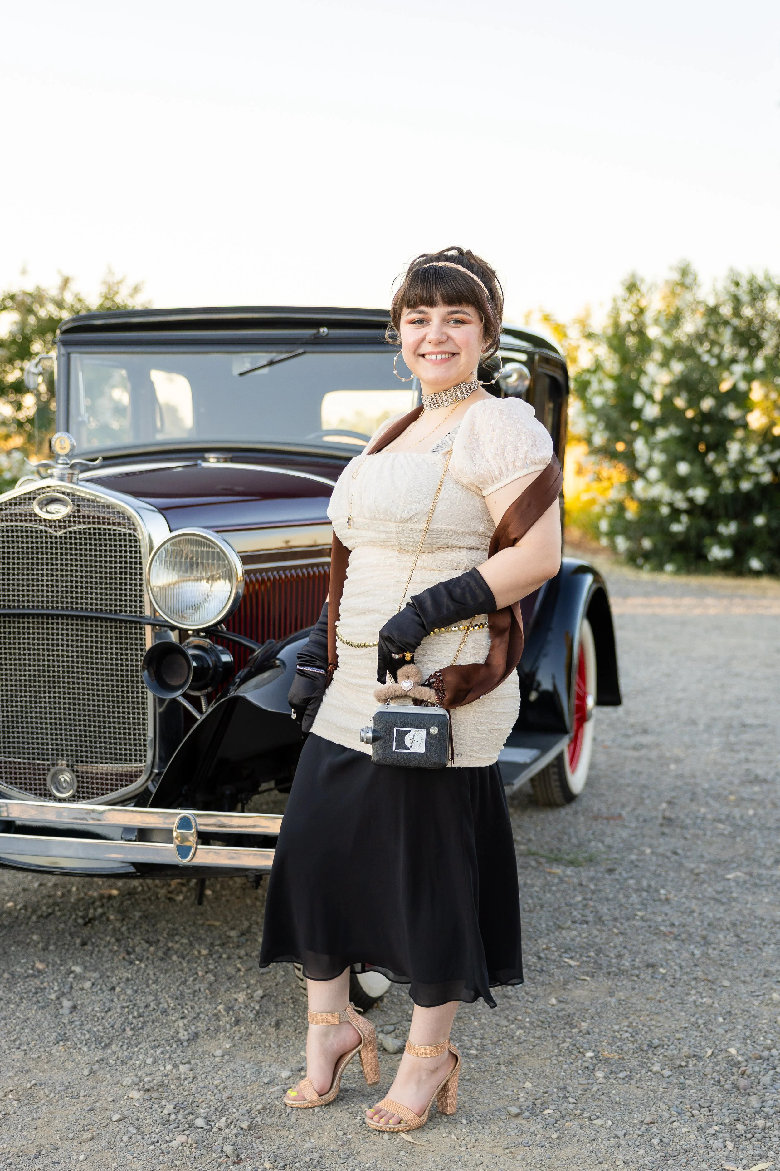 Young woman dressed in vintage style standing in front of a black classic car outdoors during daytime, wearing a cream-colored dress, black gloves, and high heels.