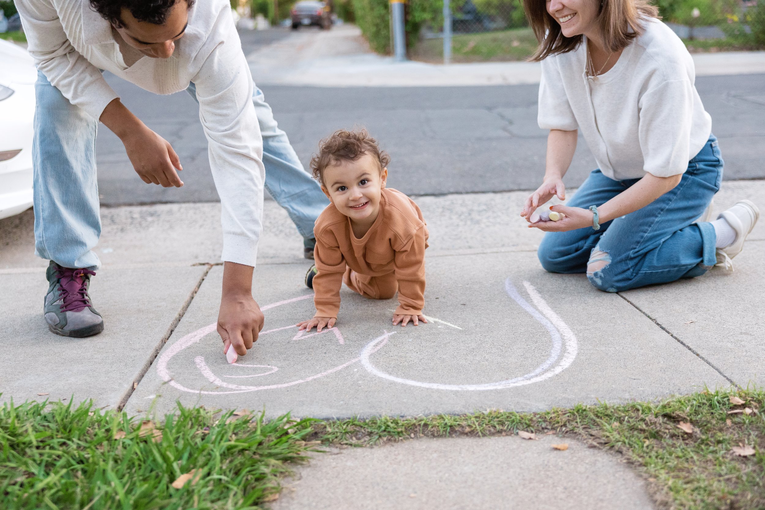 A man, a young woman, and a small child drawing with chalk on a concrete sidewalk in a park or neighborhood. The child is in the center, crawling and smiling at the camera. The woman is kneeling and holding chalk, smiling, while the man is bent over,