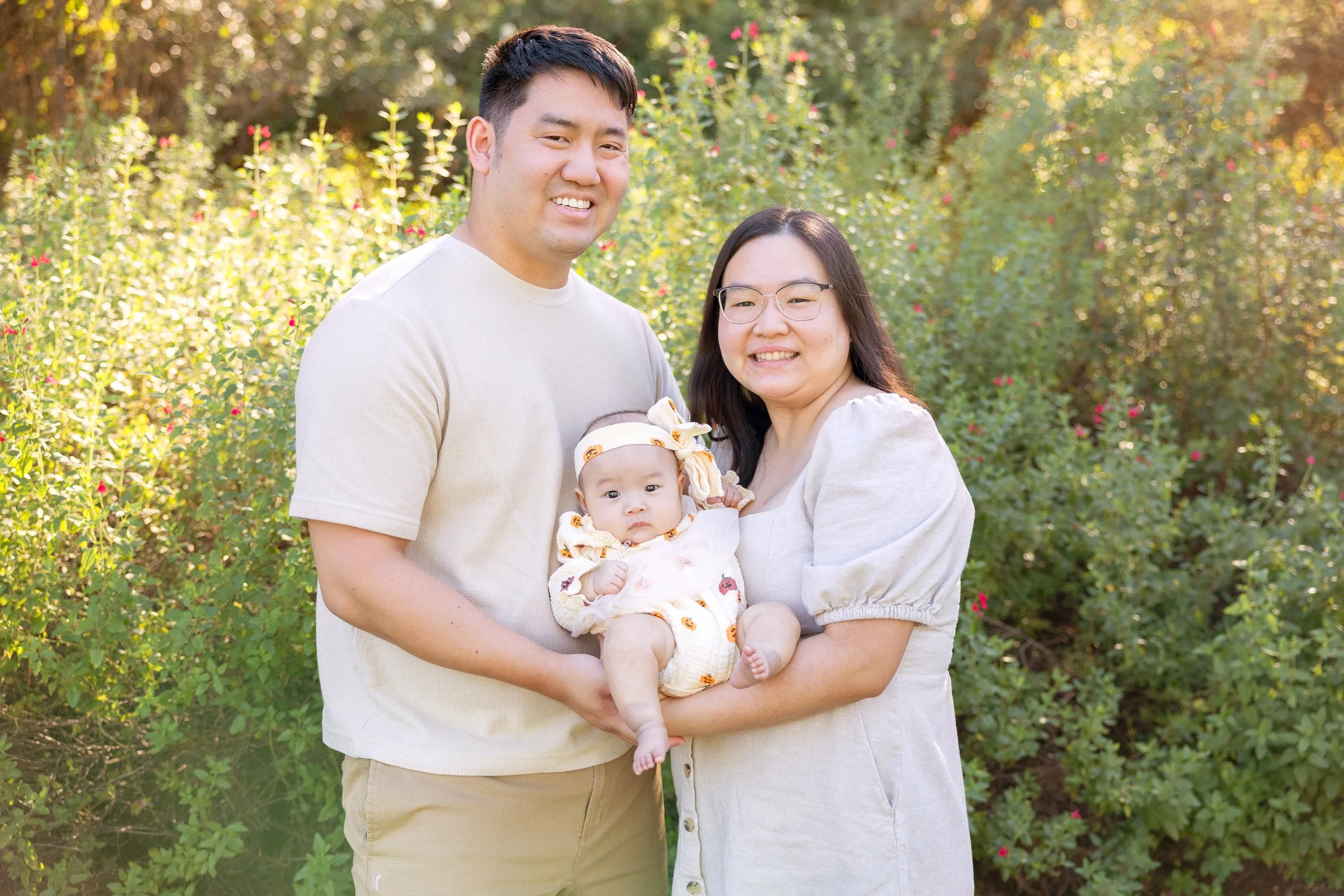 A young couple stands together outdoors, smiling while holding their baby dressed in a floral outfit, surrounded by tall greenery in soft golden light.