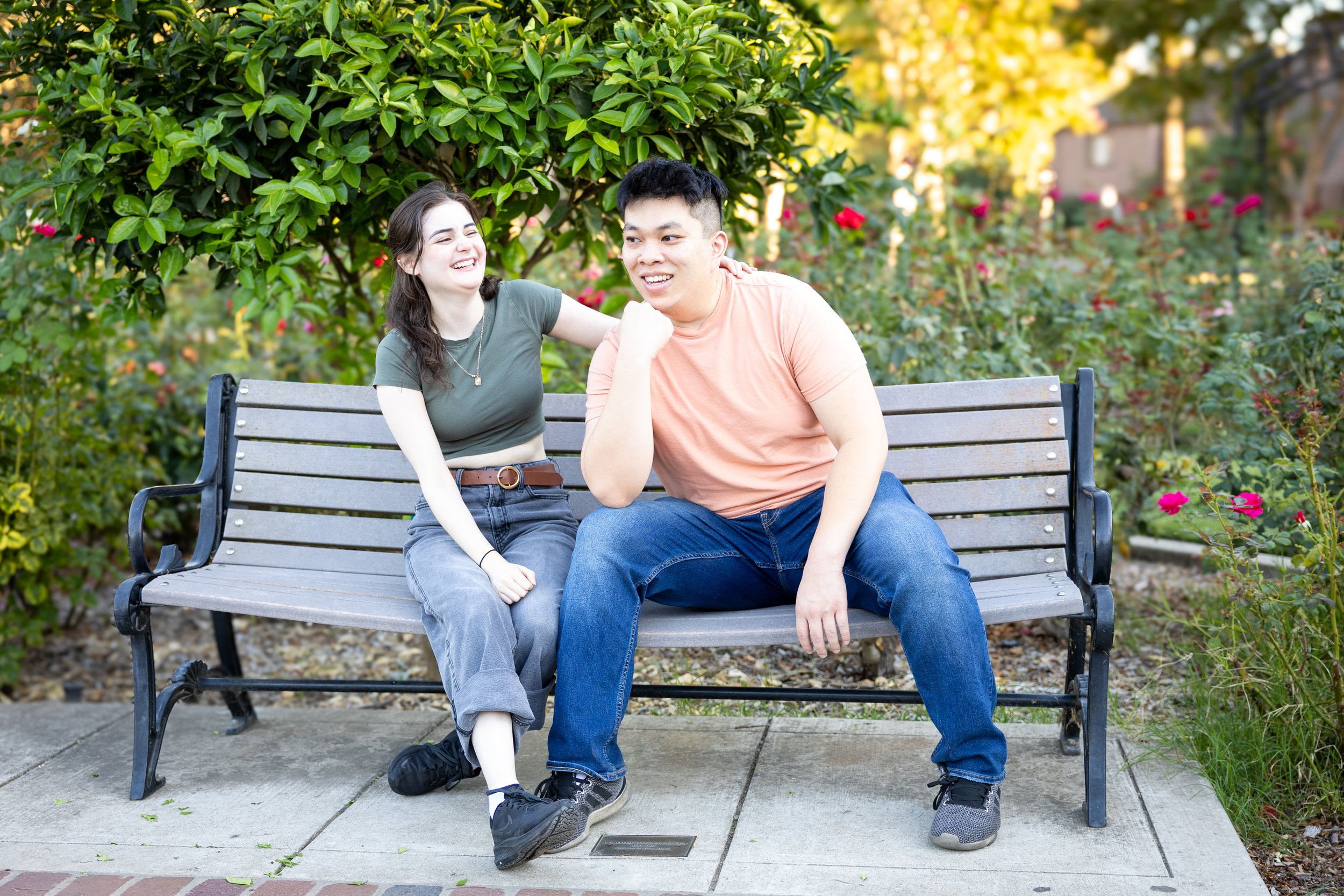 A young woman and a young man sitting on a park bench, smiling and laughing, with greenery and flowers in the background.
