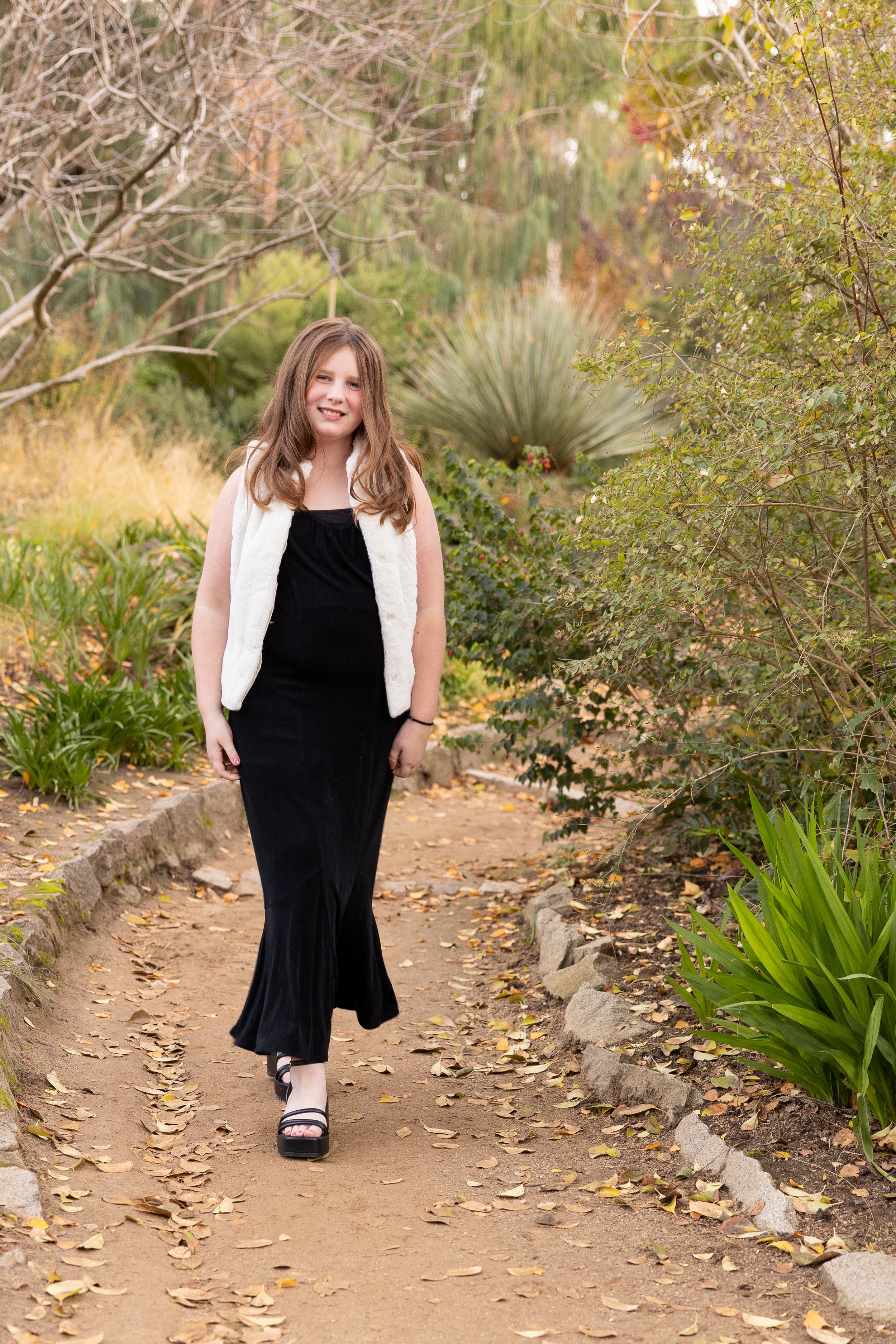 A young girl with long brown hair, wearing a black dress and white vest, walking on a leaf-covered dirt path in a garden with green plants and trees, during autumn.