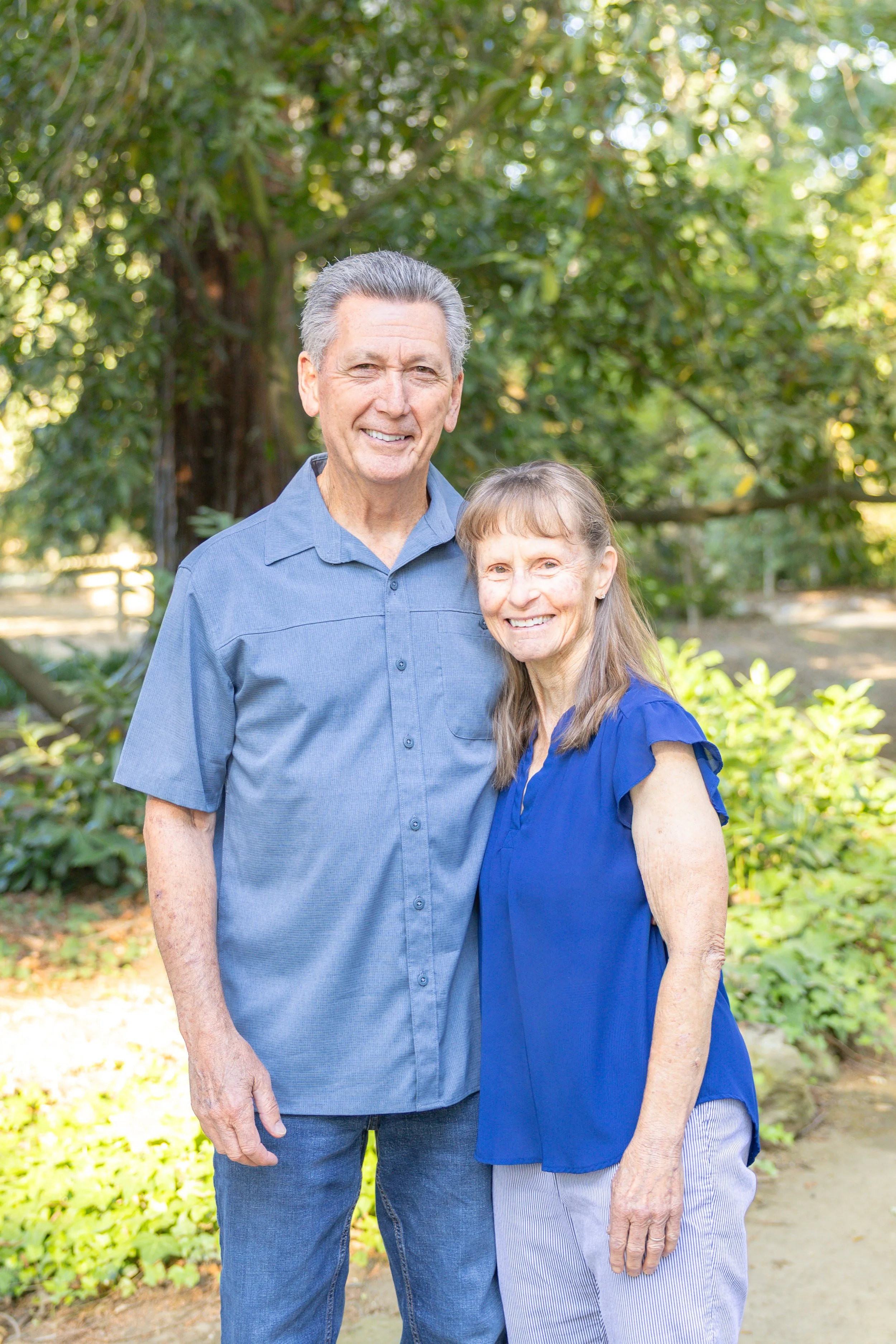 A man and woman stand closely together, smiling at the camera in a shaded park surrounded by trees and greenery.