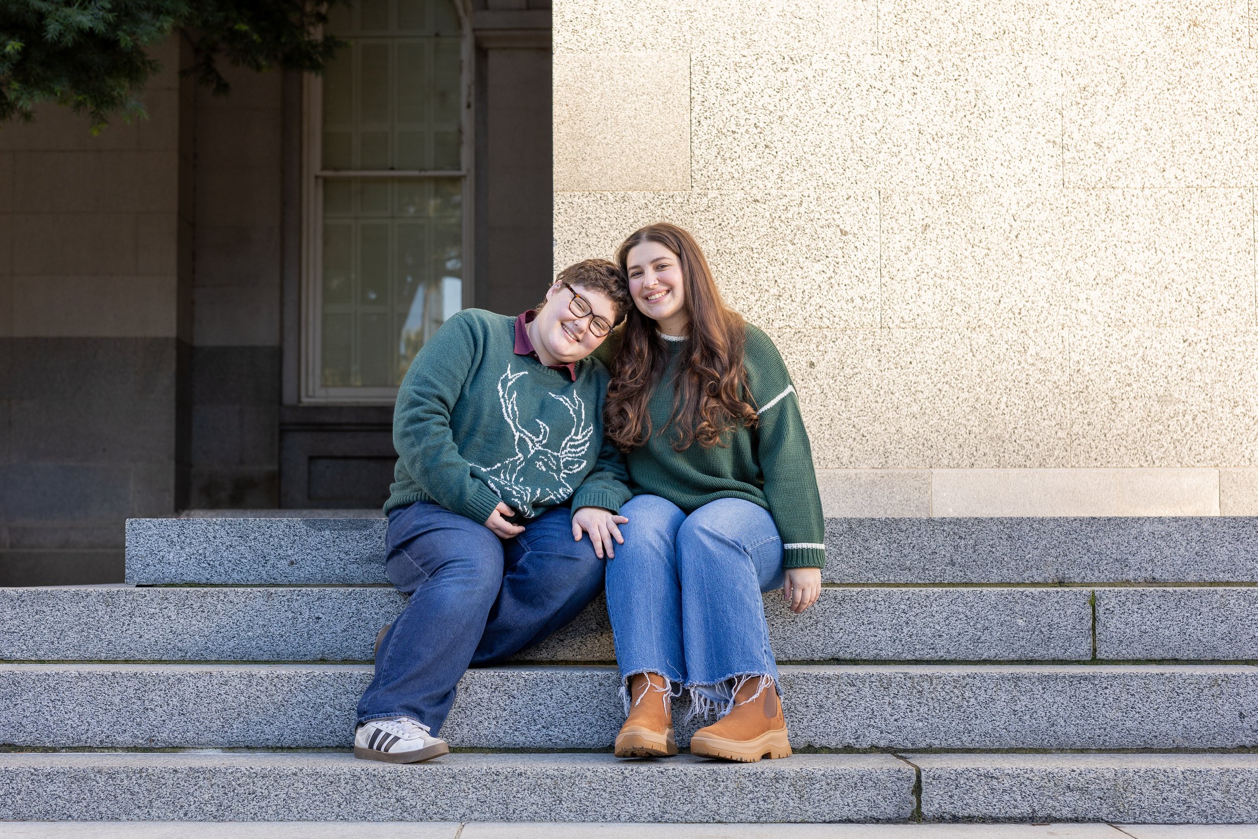 Two young women with sitting on stone steps outside, smiling and leaning their heads together.