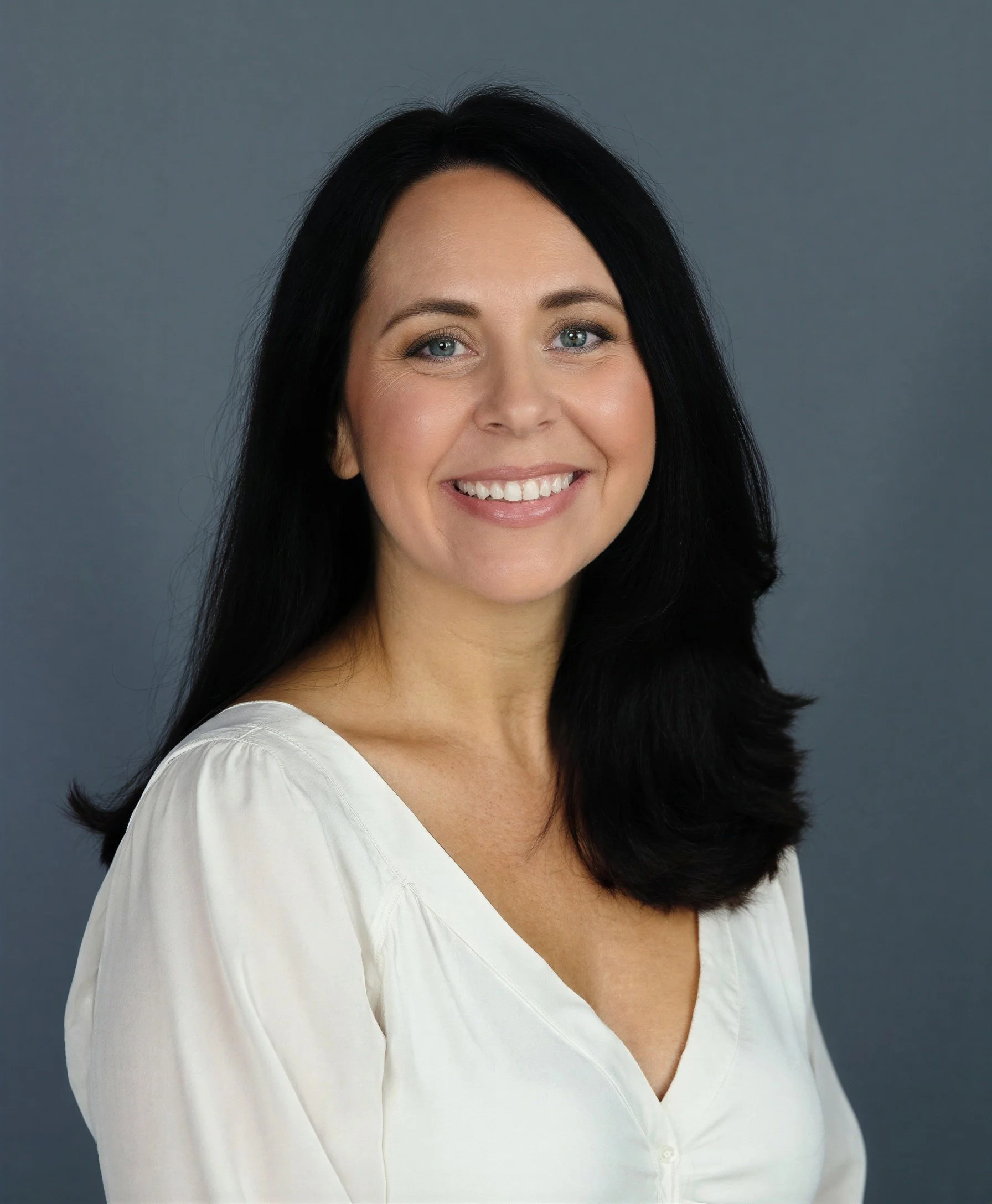 Close-up portrait of a smiling woman with long black hair, blue eyes, wearing a white V-neck top, against a gray background.