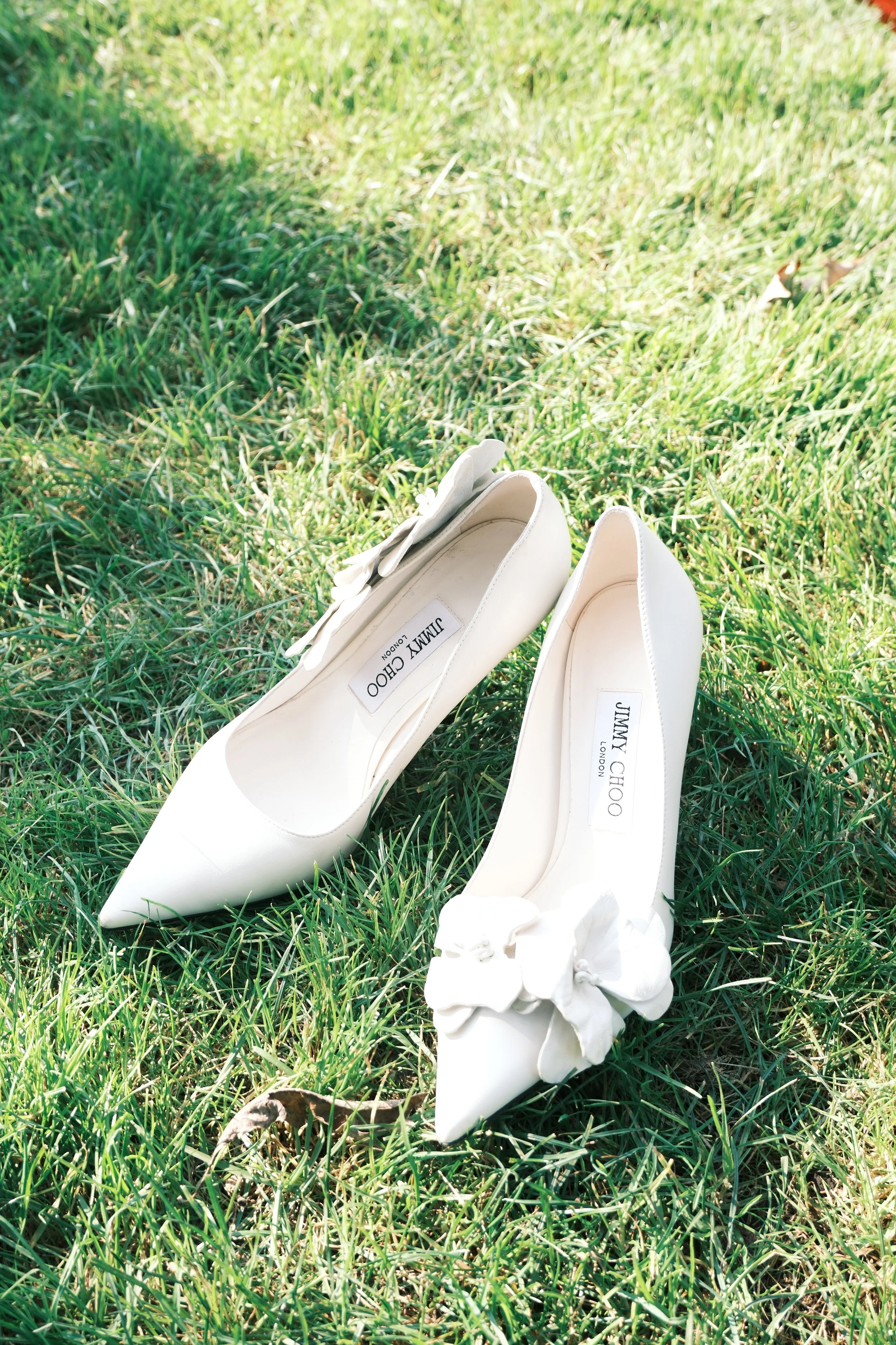 White high-heeled shoes with floral embellishments on green grass.