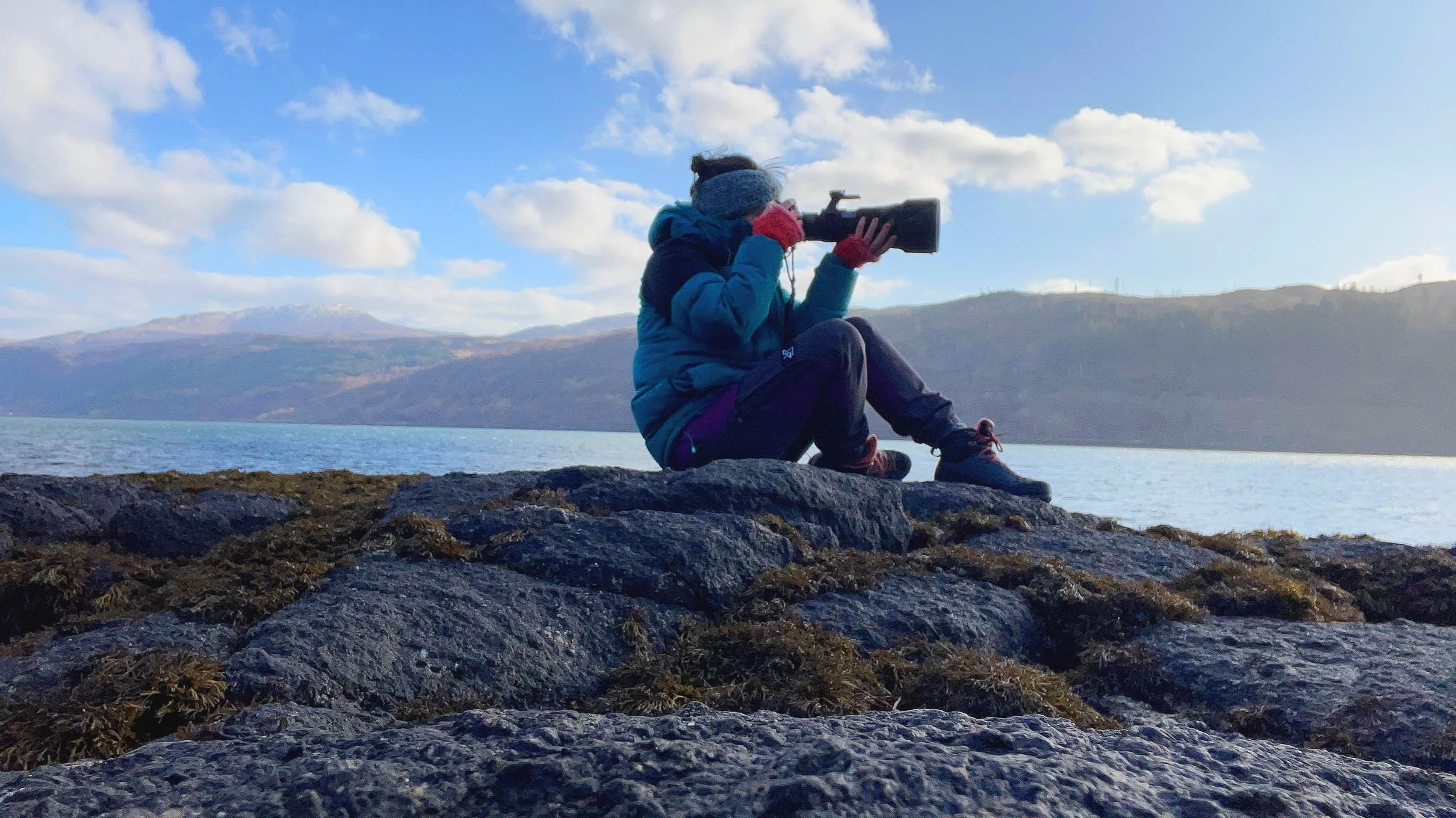 Person sitting on rocks by a lake, taking photos with a camera. They are wearing a blue jacket, gray hat, red gloves, and black pants, with a backdrop of mountains, water, and a partly cloudy sky.