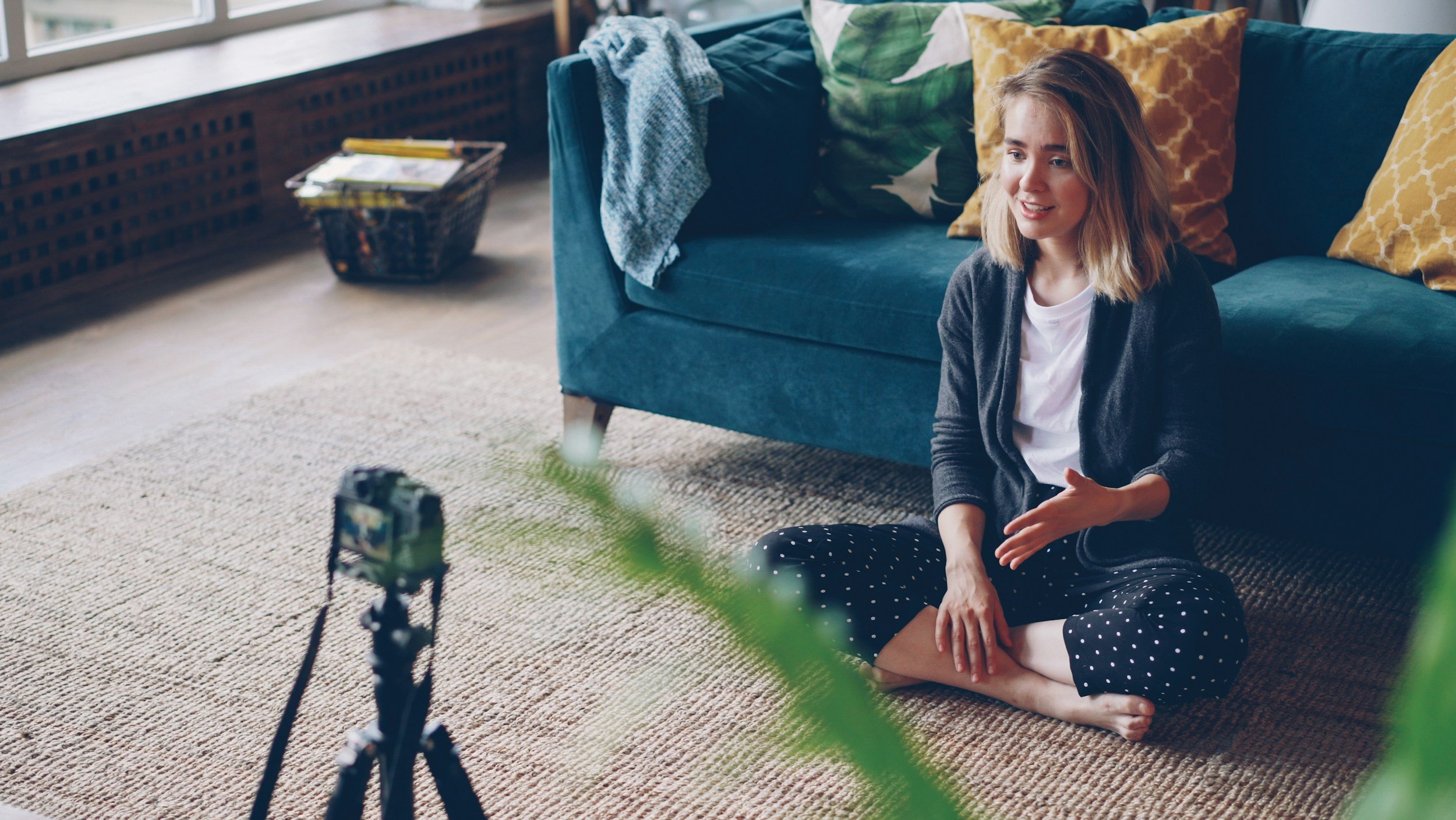 A young woman sitting on a carpeted floor in a living room, being filmed by a camera on a tripod. She has shoulder-length blonde hair, is smiling, and is wearing a white shirt with a dark cardigan and polka dot pants. The room features a teal sofa with colorful pillows, a basket with items, and a large window with a radiator below.