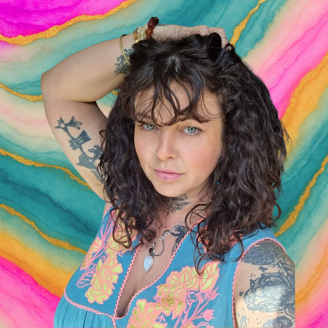 Alternative woman with curly dark hair, tattoos, and a nose and lip piercing, taking a selfie against a vibrant, colorful watercolor background. Holistic Thrive Founder and CEO.