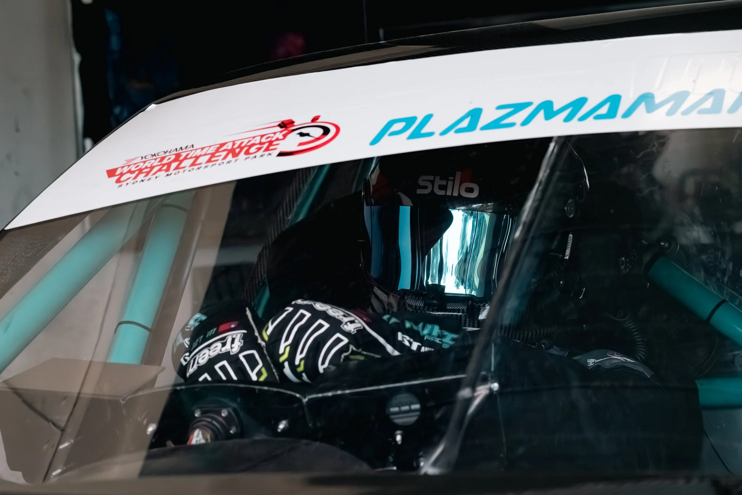 A race car driver wearing a black helmet and racing gloves sits inside a race car with a clear canopy, surrounded by sponsorship stickers.